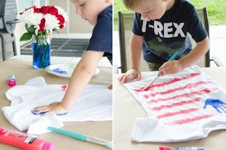 A child putting a blue paint handprint onto a white t-shirt and painting red stripes to make the shirt look like an American flag.