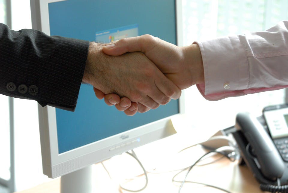 Two people shaking hands across an office desk with a computer monitor and phone in the background.