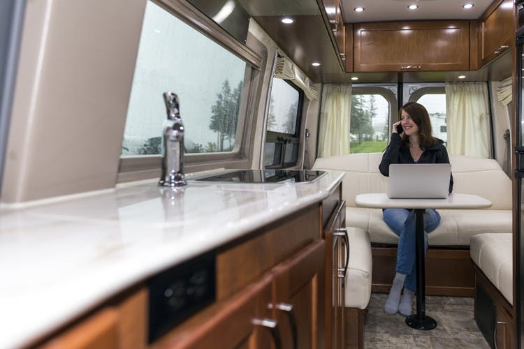 Woman inside an RV, with a laptop computer open on the table, talking on the phone.