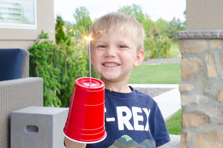 A child holding up a sparkler that has been lit and fed through a hole in a plastic cup that is covering the child's hand.