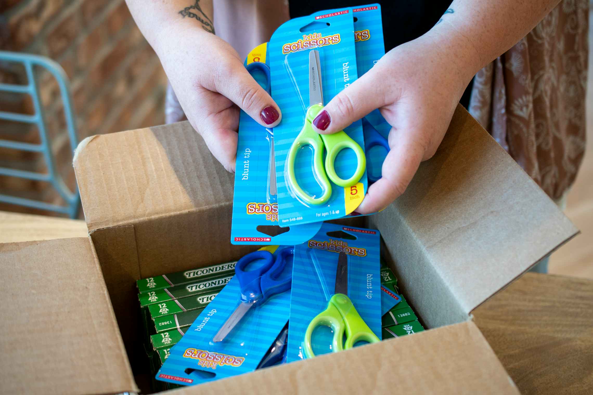 A person taking multiple pairs of scissors out of a shipping box with more scissors and boxes of pencils in it.
