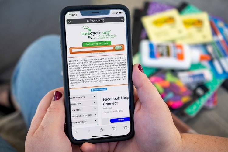 A person's hands holding up an iPhone, displaying the freecycle.org website, in front of a pile of school supplies.
