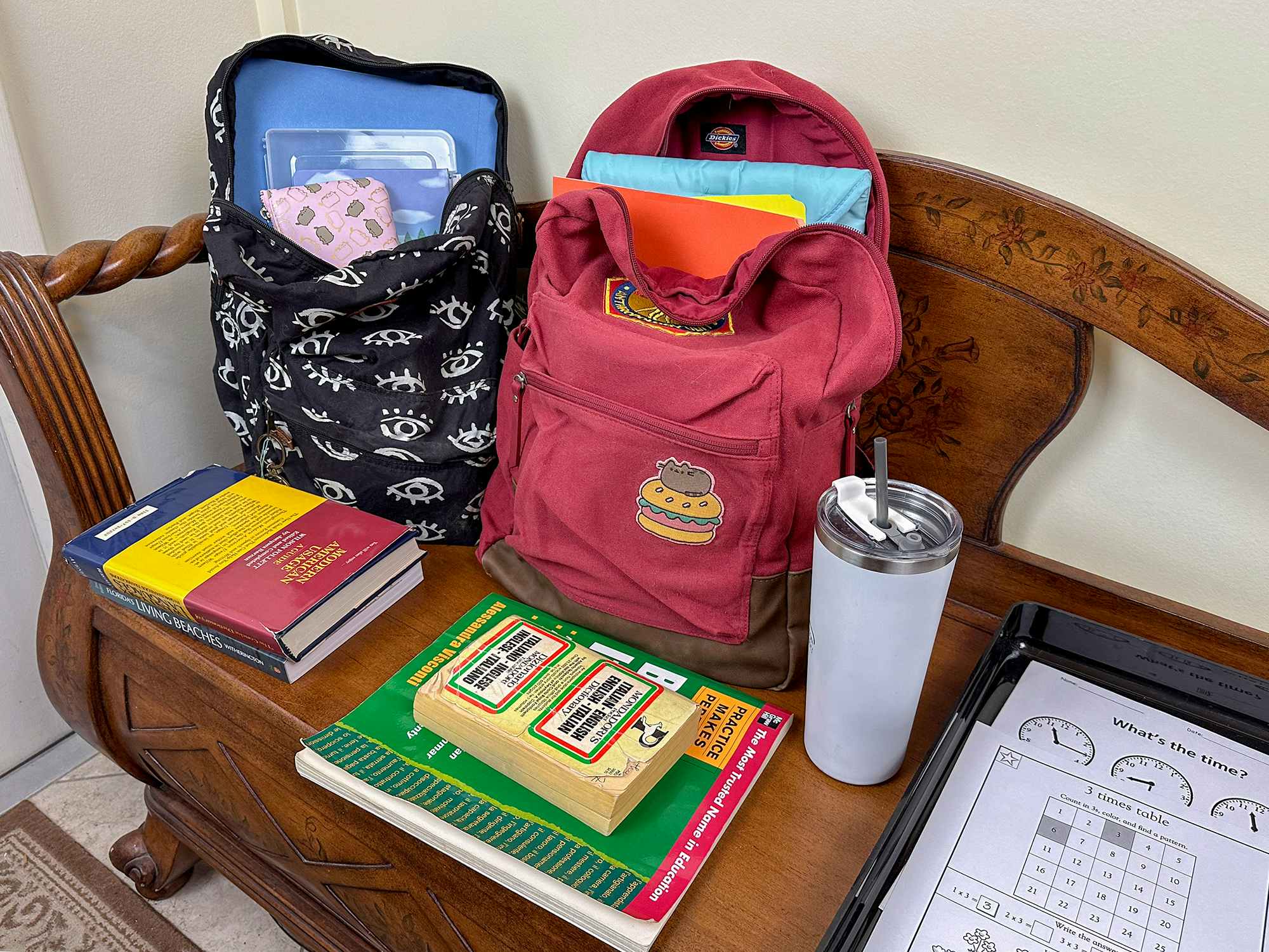 Backpacks, books, a water bottle, and a tray for assignments set up on a wooden bench by the door