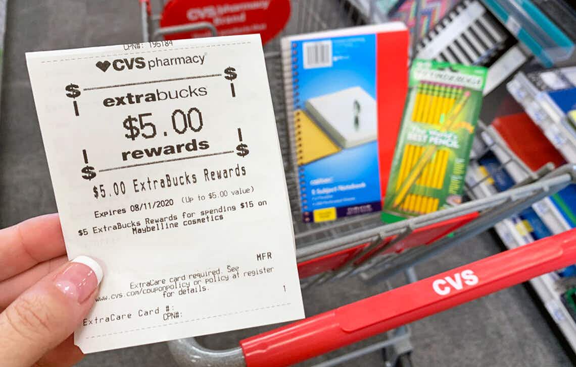 A person's hand holding CVS receipt with $5 extra bucks rewards above a CVS shopping cart with school supplies in the basket.