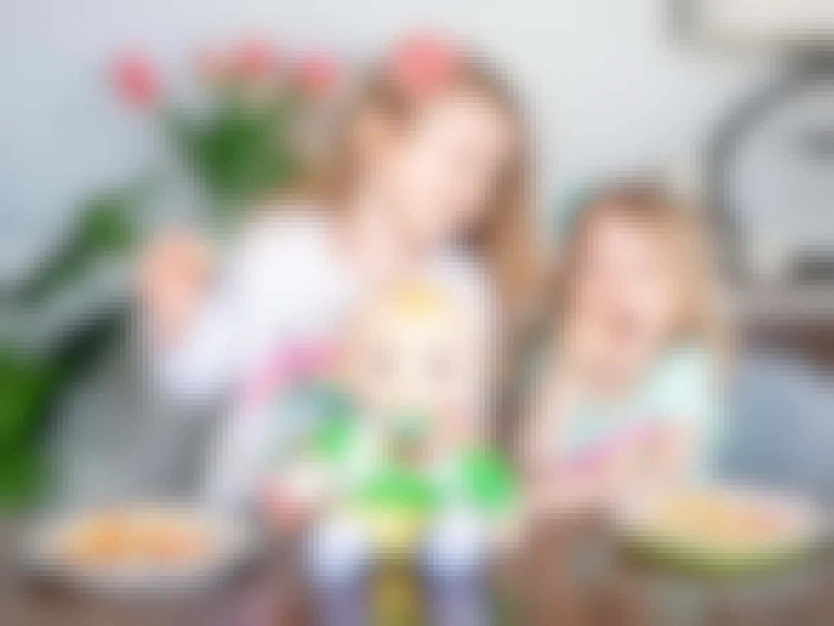 Two kids at a table in a dinning room, eating sliced apples with a Cocomelon doll between them.