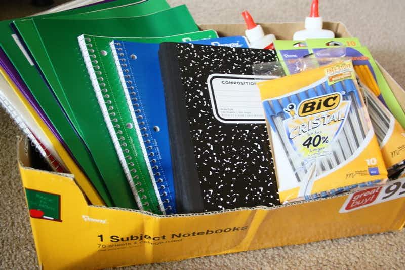 School supplies, including folders, notebooks, pens, and glue sitting in a box on a carpeted floor.