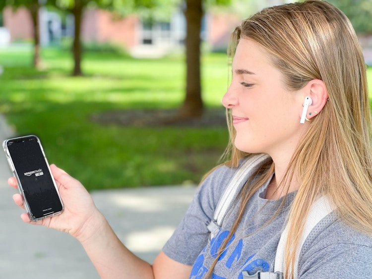 A college student with Airpods in her ears and the Amazon Music app start screen displayed on her phone.