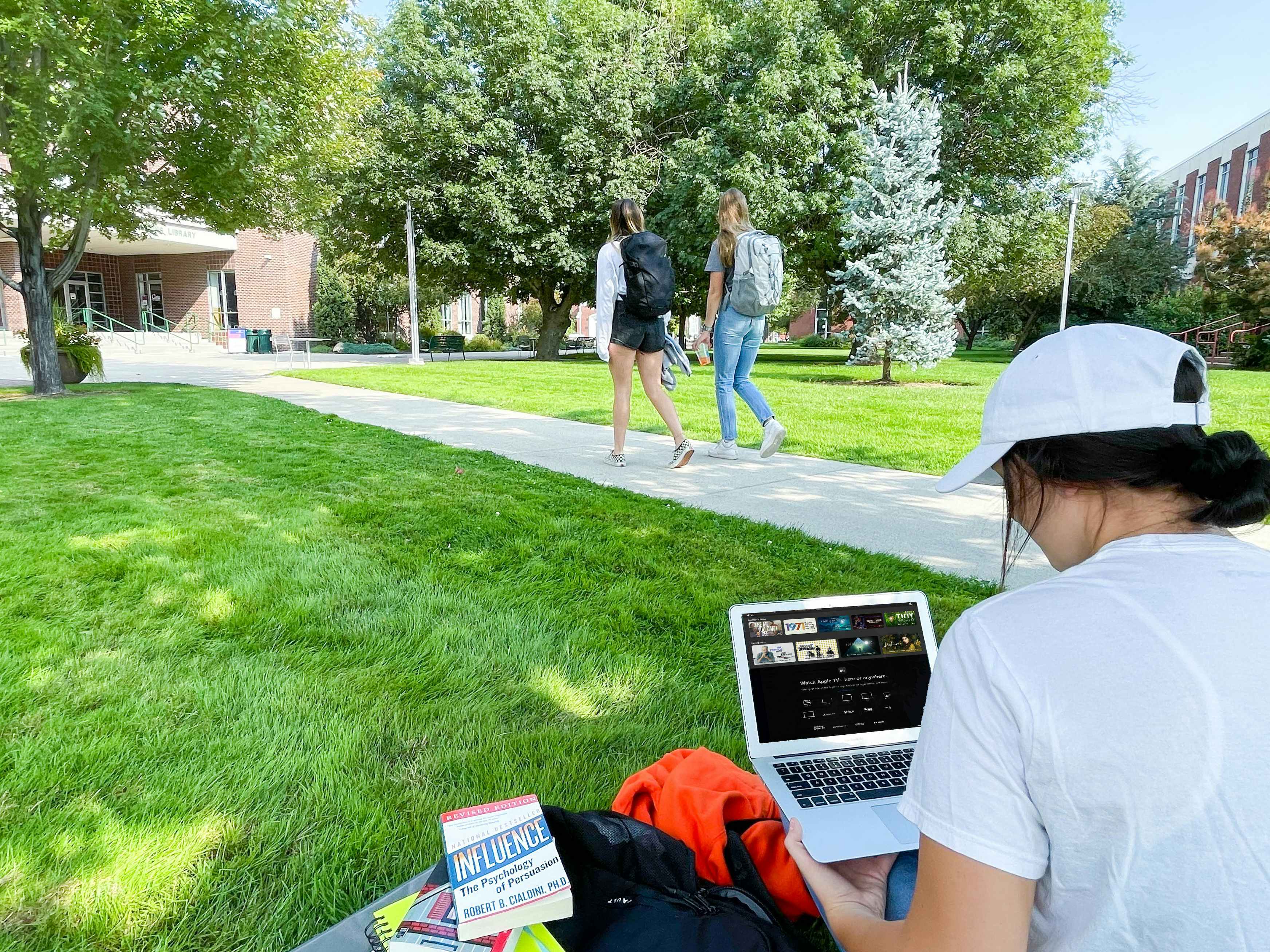 A college student sitting in grass on campus and watching Apple TV on laptop with two more students walking by on the sidewalk in the background.