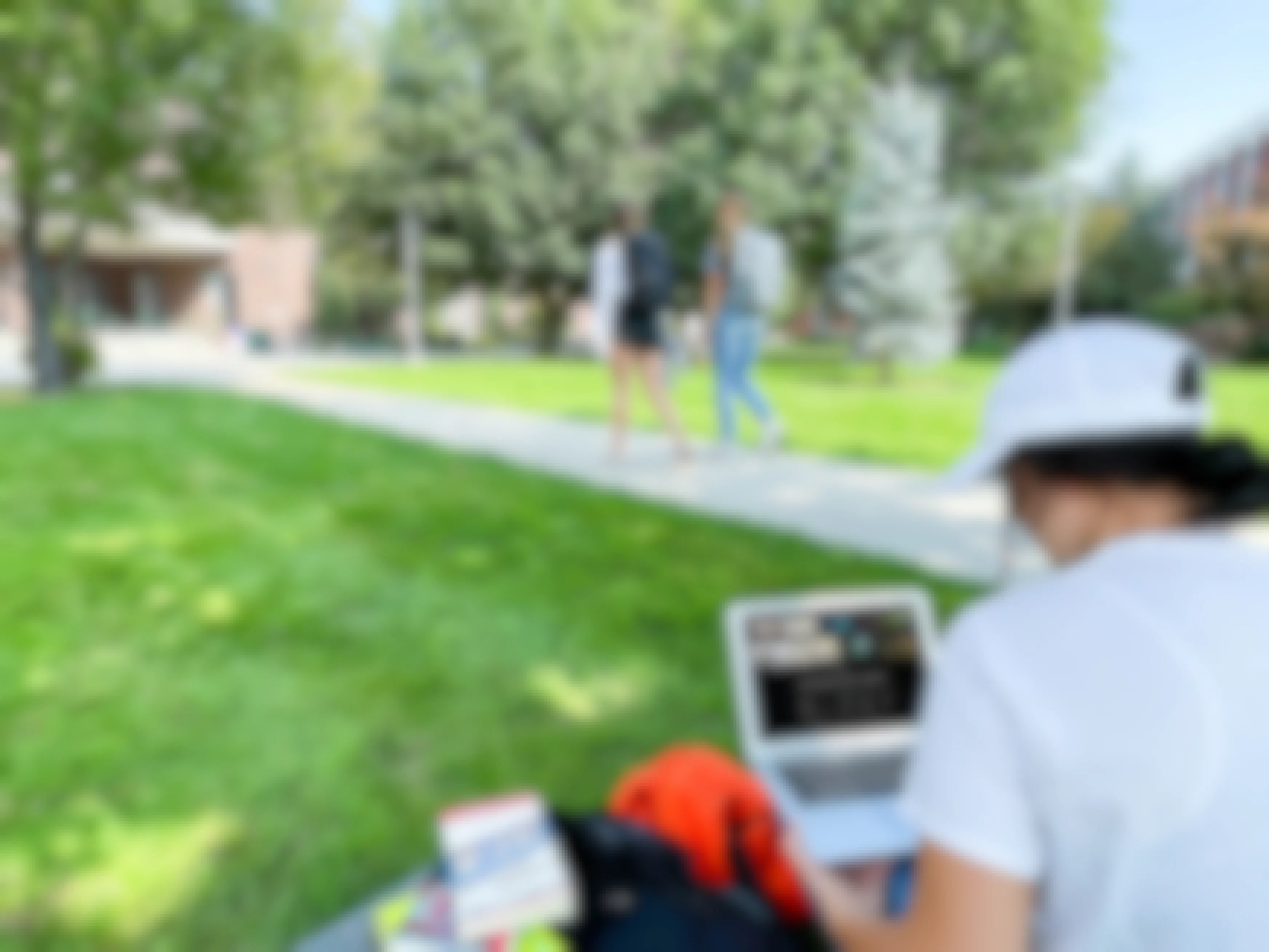 A college student sitting in grass on campus and watching Apple TV on laptop with two more students walking by on the sidewalk in the background.