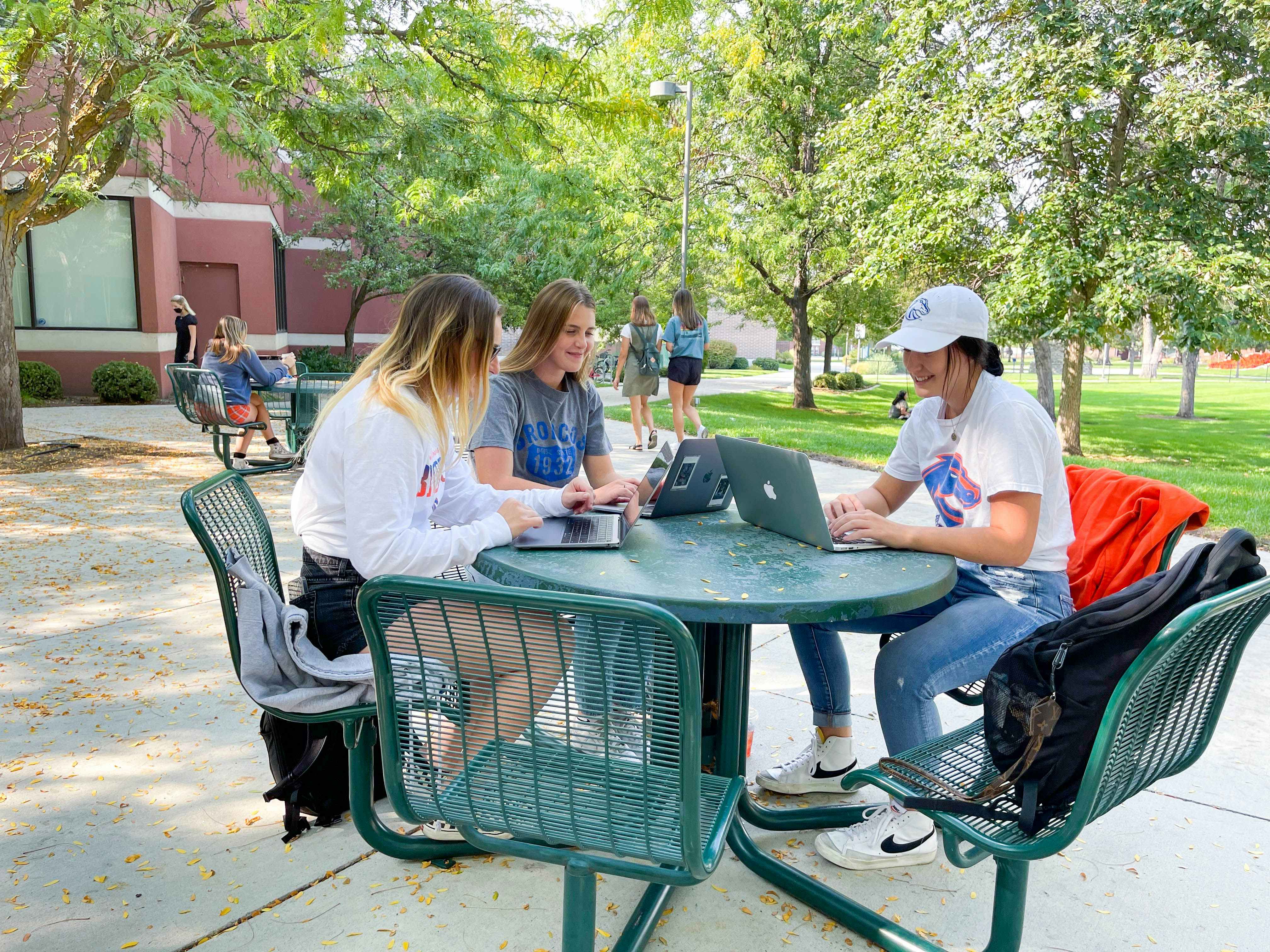 - Three college students are sitting outside at a green table on campus. They are all looking at their laptops and talking.