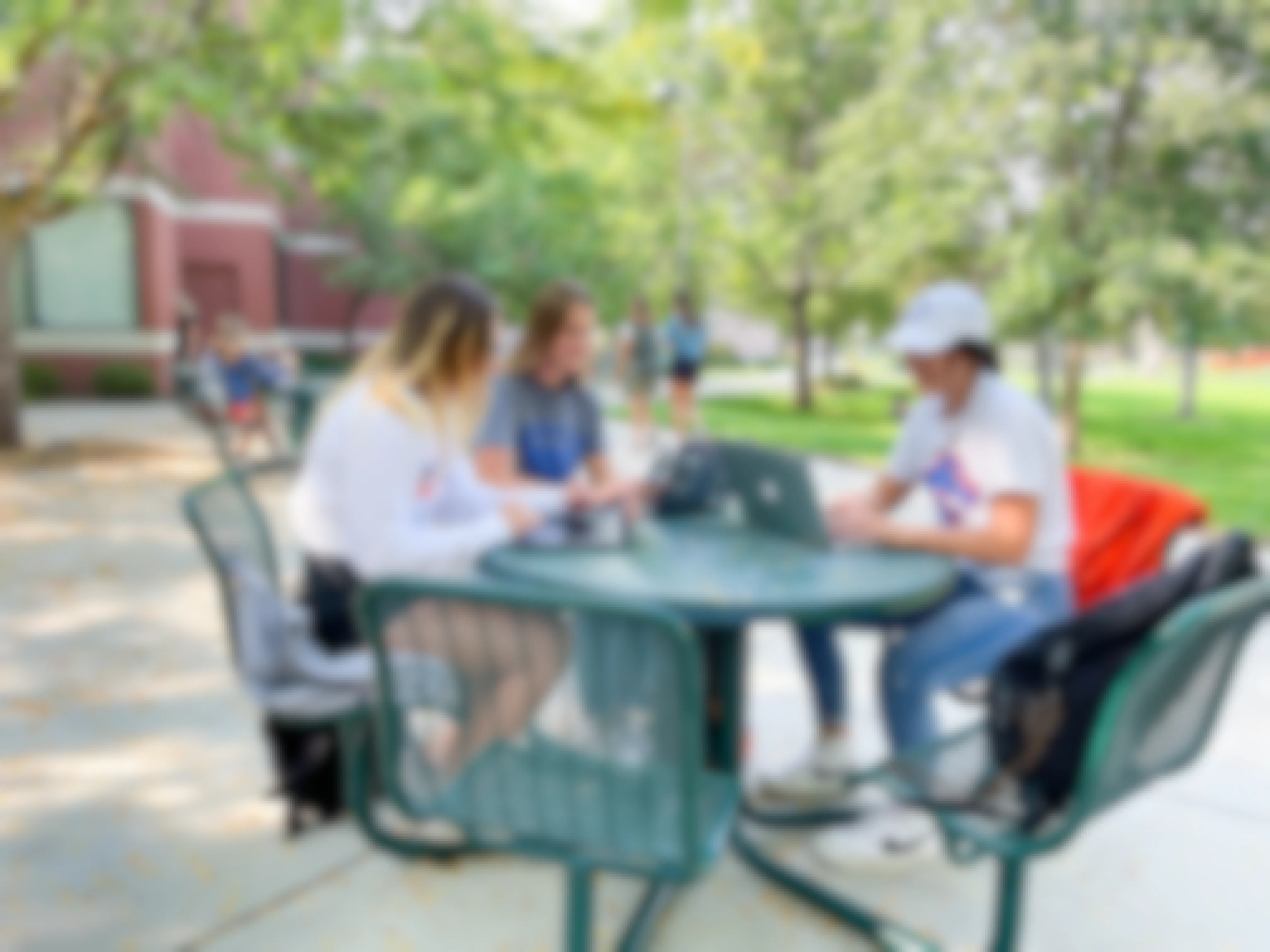 - Three college students are sitting outside at a green table on campus. They are all looking at their laptops and talking.