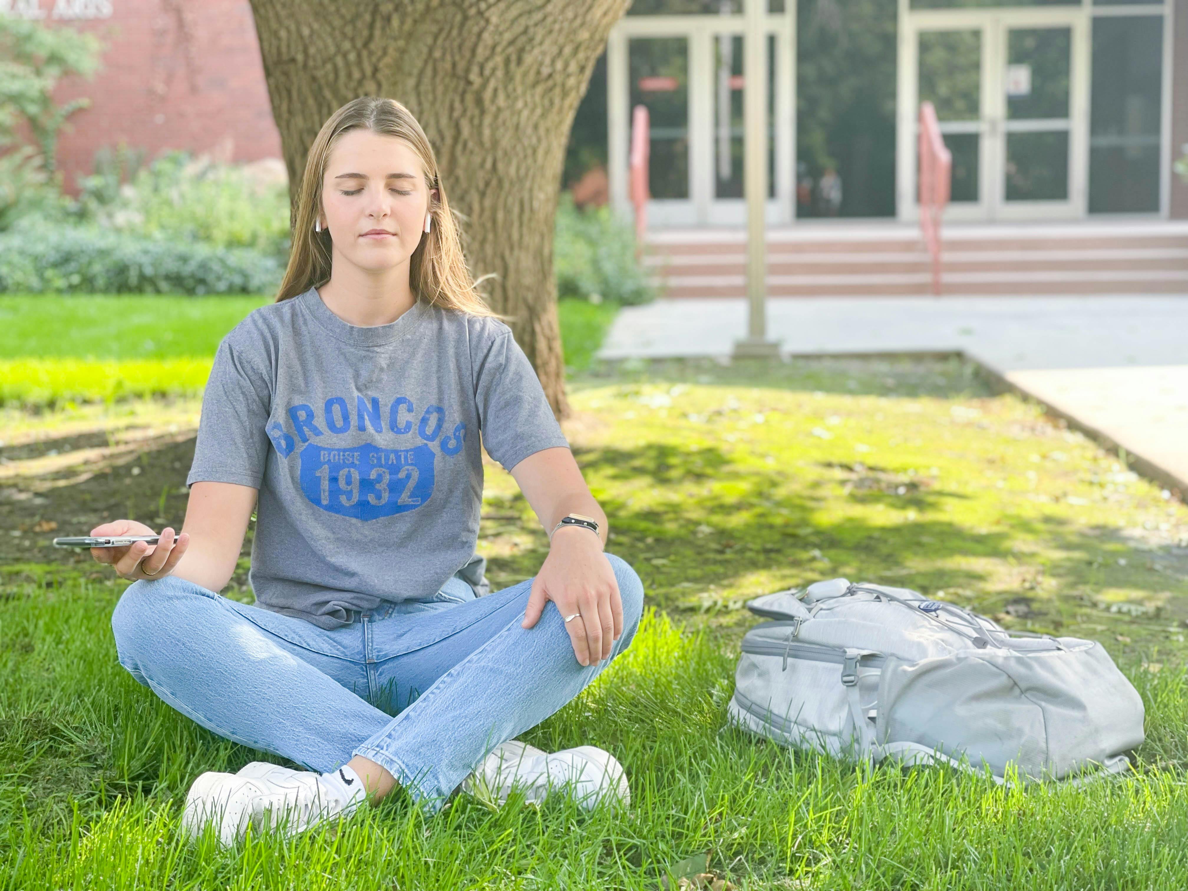 A student with wireless earbuds sitting under a tree, meditating with a phone in her hand.