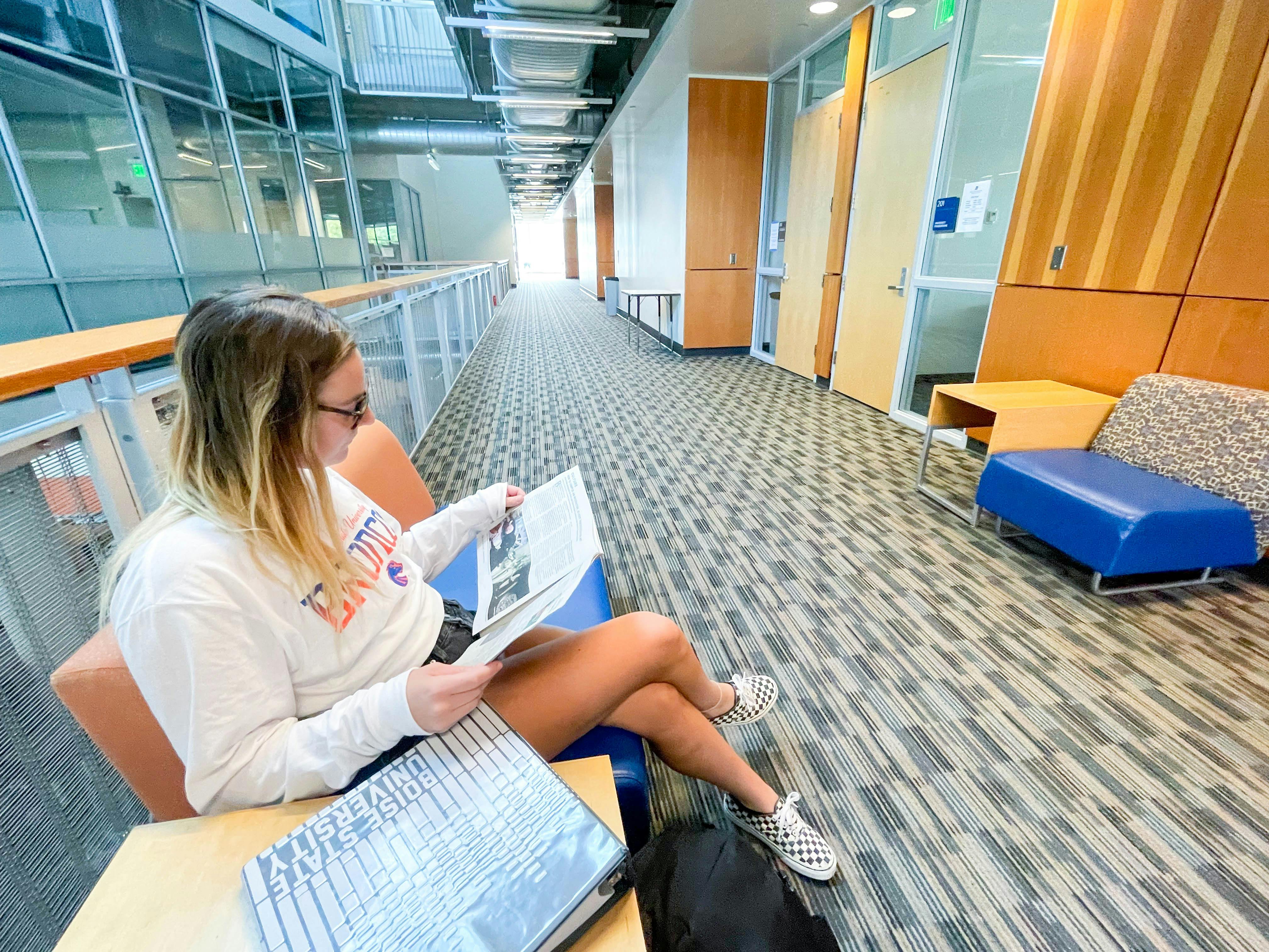 A college student sitting outside of a classroom reading a newspaper.