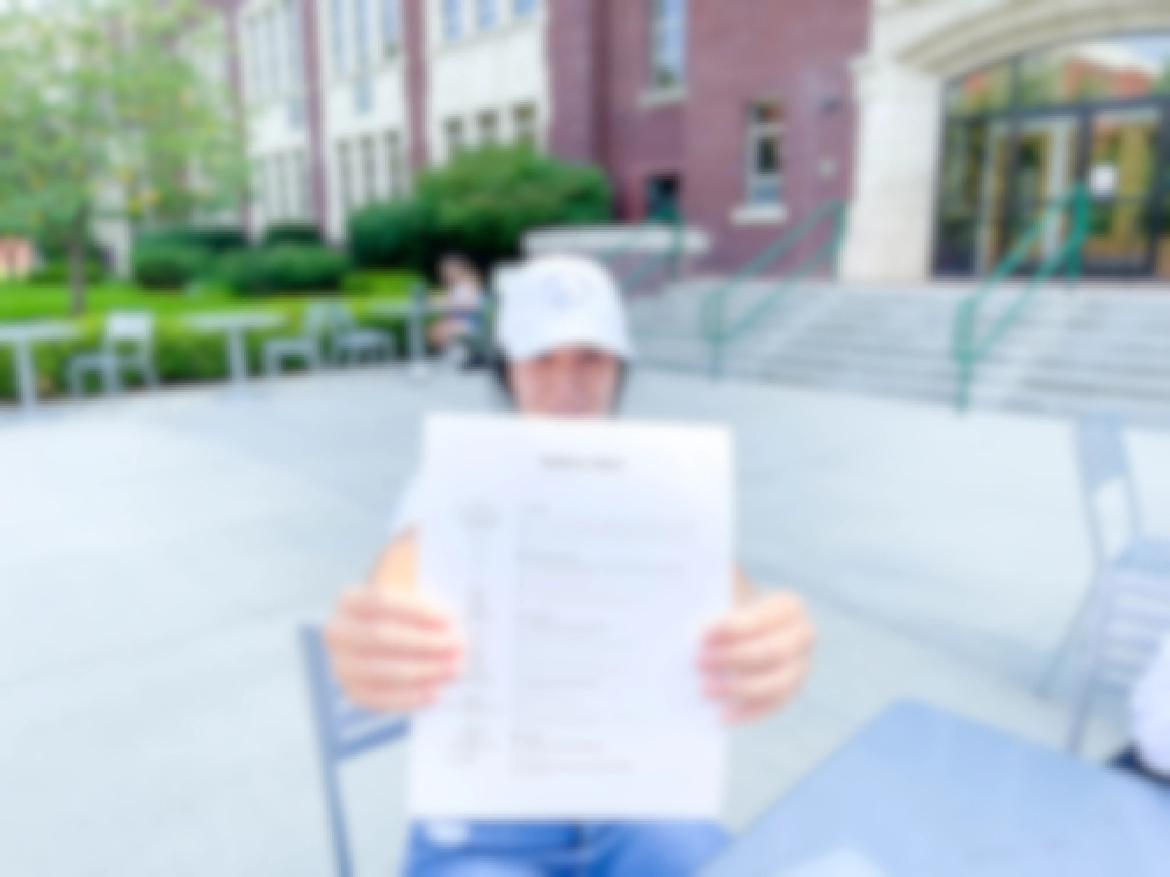 A college student sitting outside of a building on campus and holding a resume up to the camera.