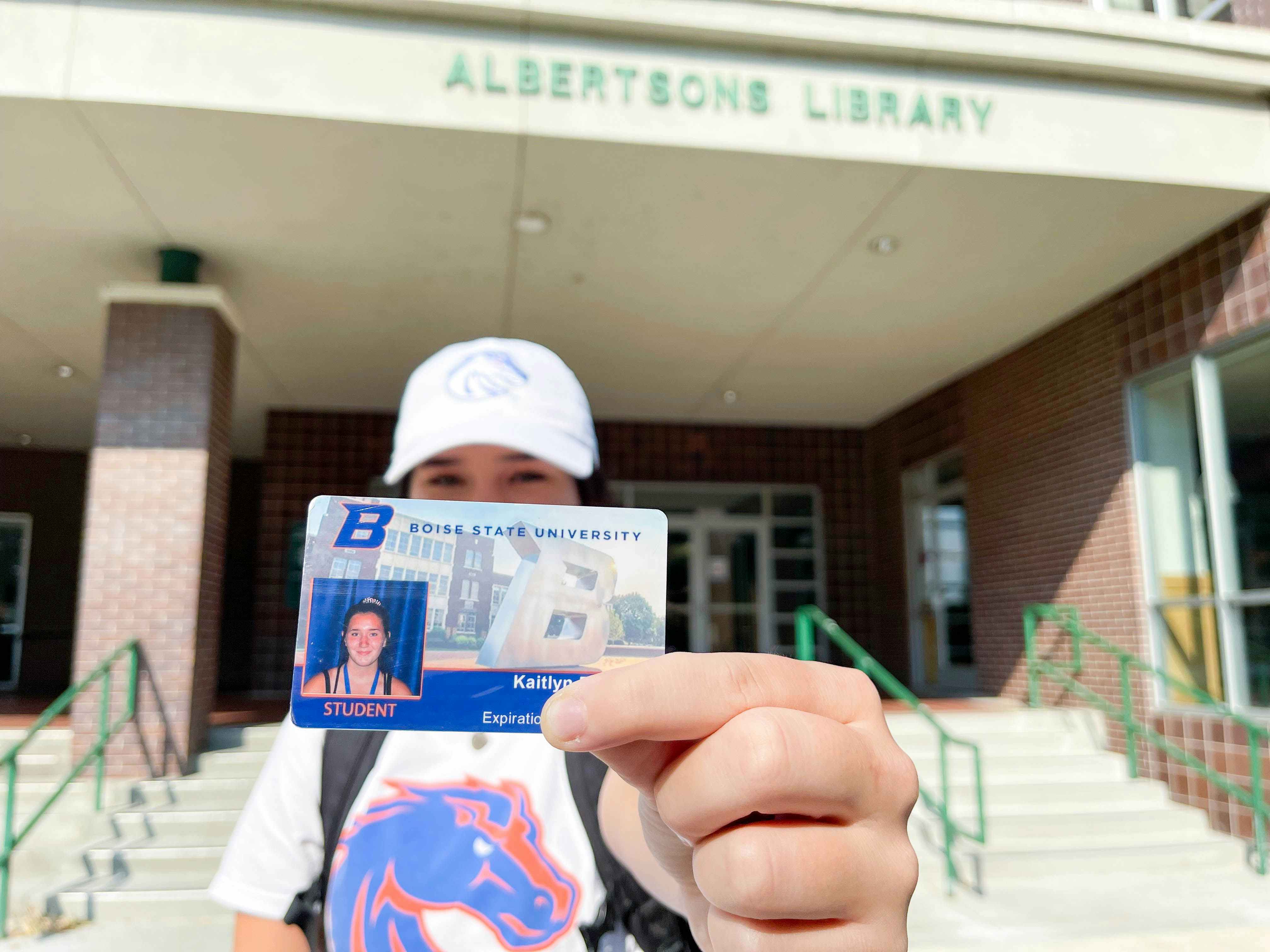 A college student holding out their student ID in front of an on-campus library.