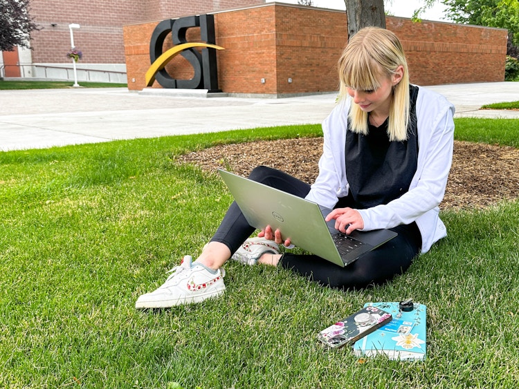 student on college campus sittiung under a tree on a laptop 