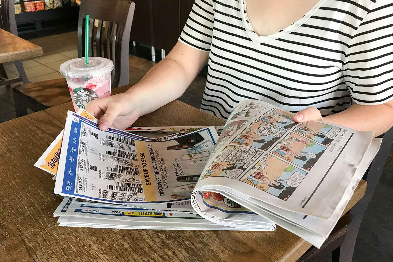 A person holding some coupons while sitting at a table with a starbucks cup on the table next to the coupons.