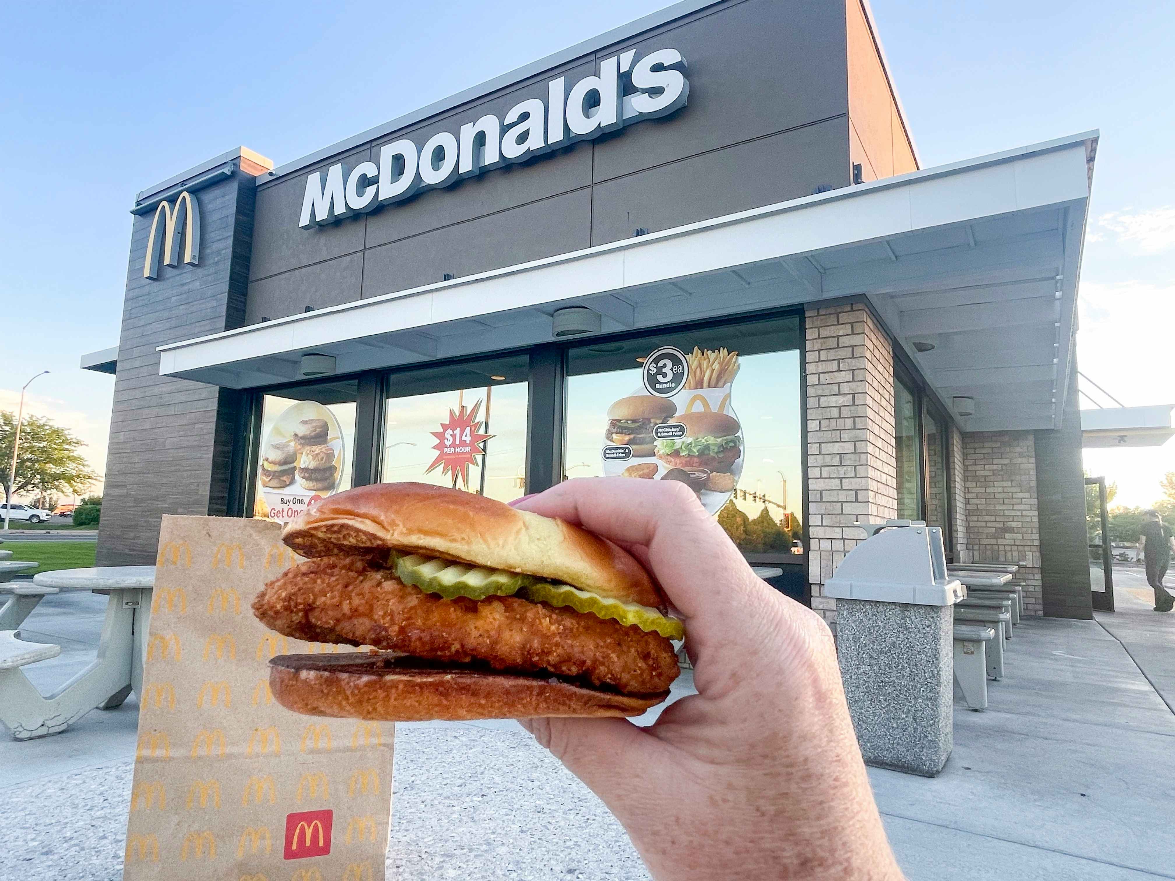 a person holding up a mcdonlds crispy chicken sandwich in front to go bag outside a mcdonalds store, the best fast food deals for chicken sandwiches