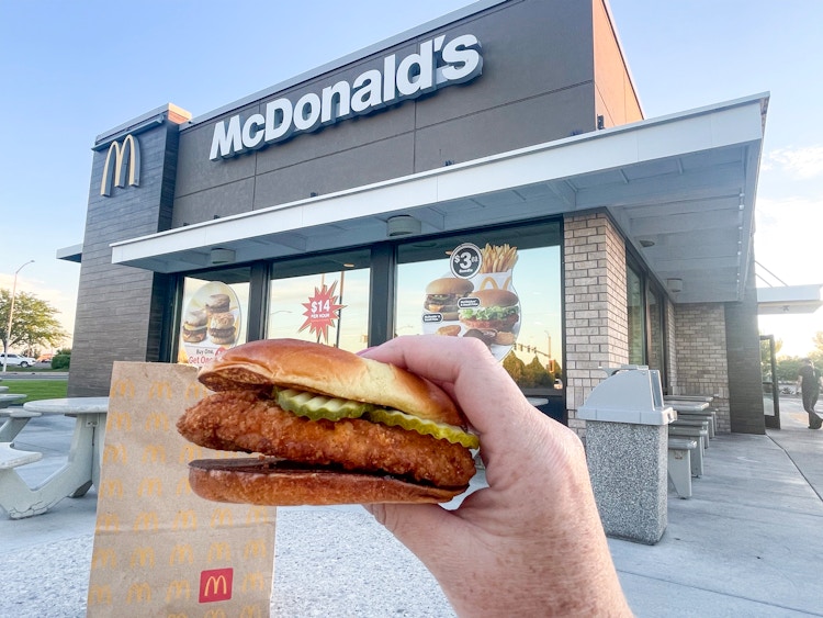 a person holding up a mcdonalds crispy chicken sandwich in front to go bag outside a mcdonalds store