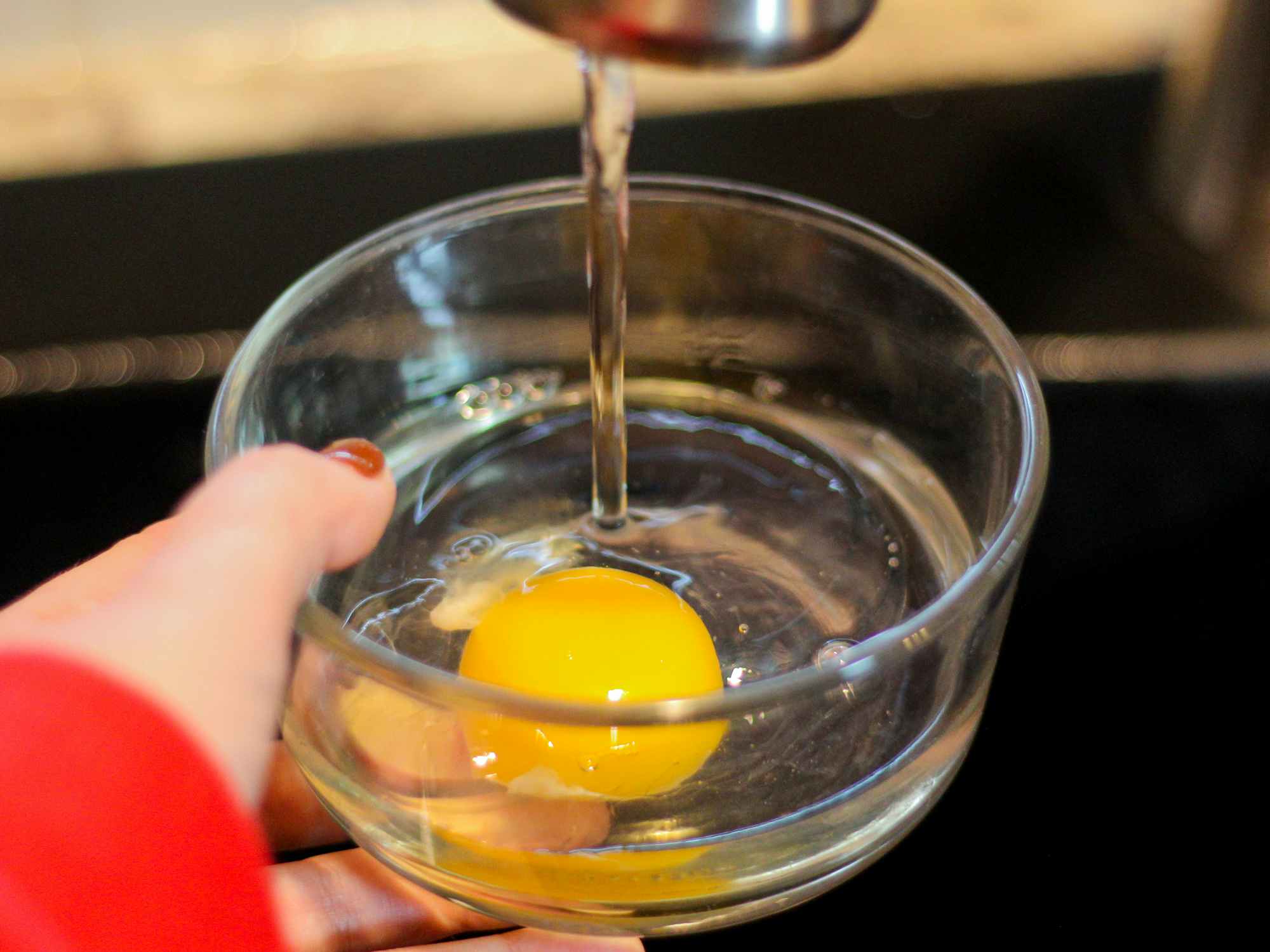 a cracked egg in a glass bowl with water from a water faucet going into the bowl