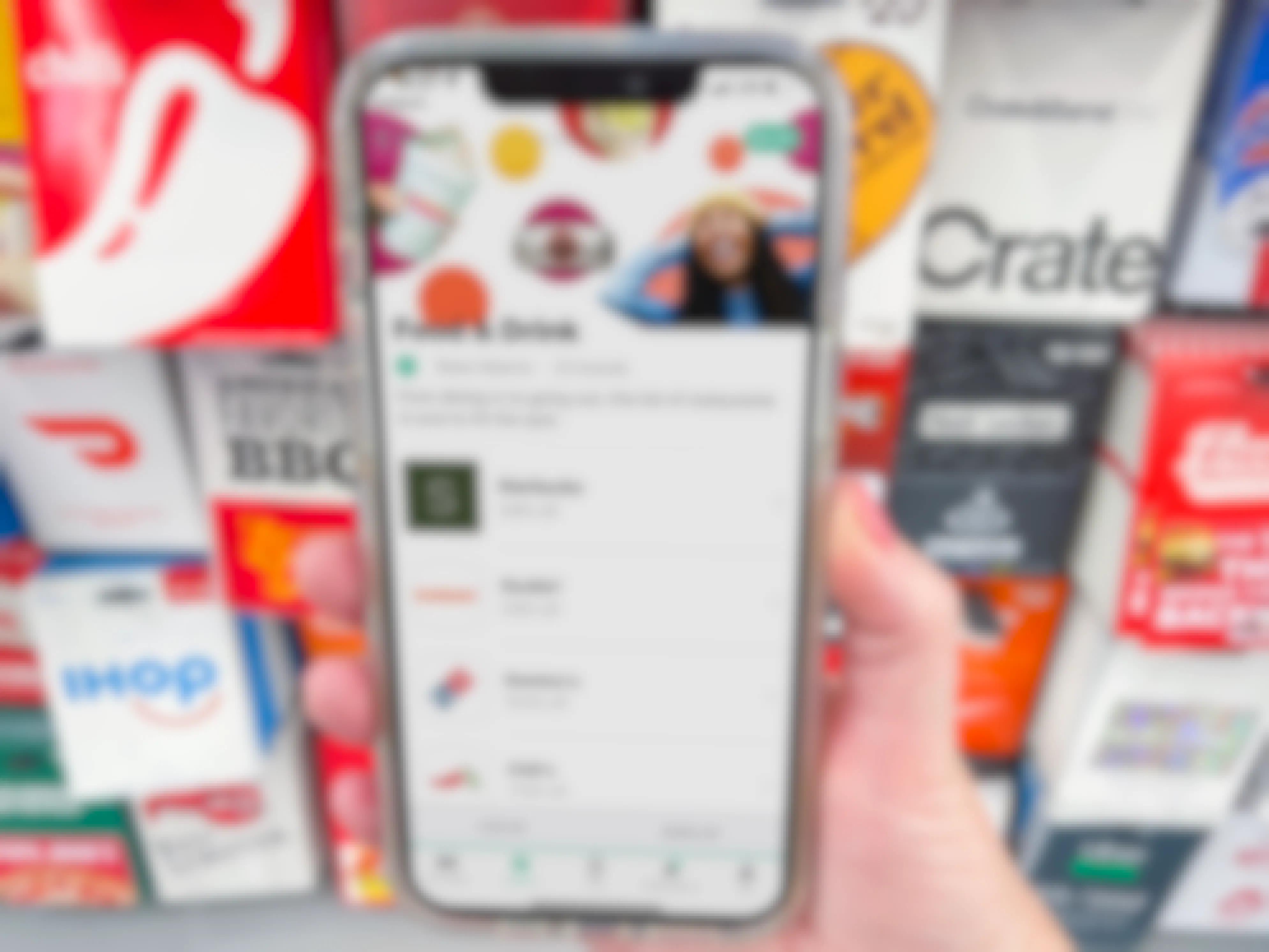 A person's hand holding an iPhone displaying the Raise app in front of a rack of gift cards.