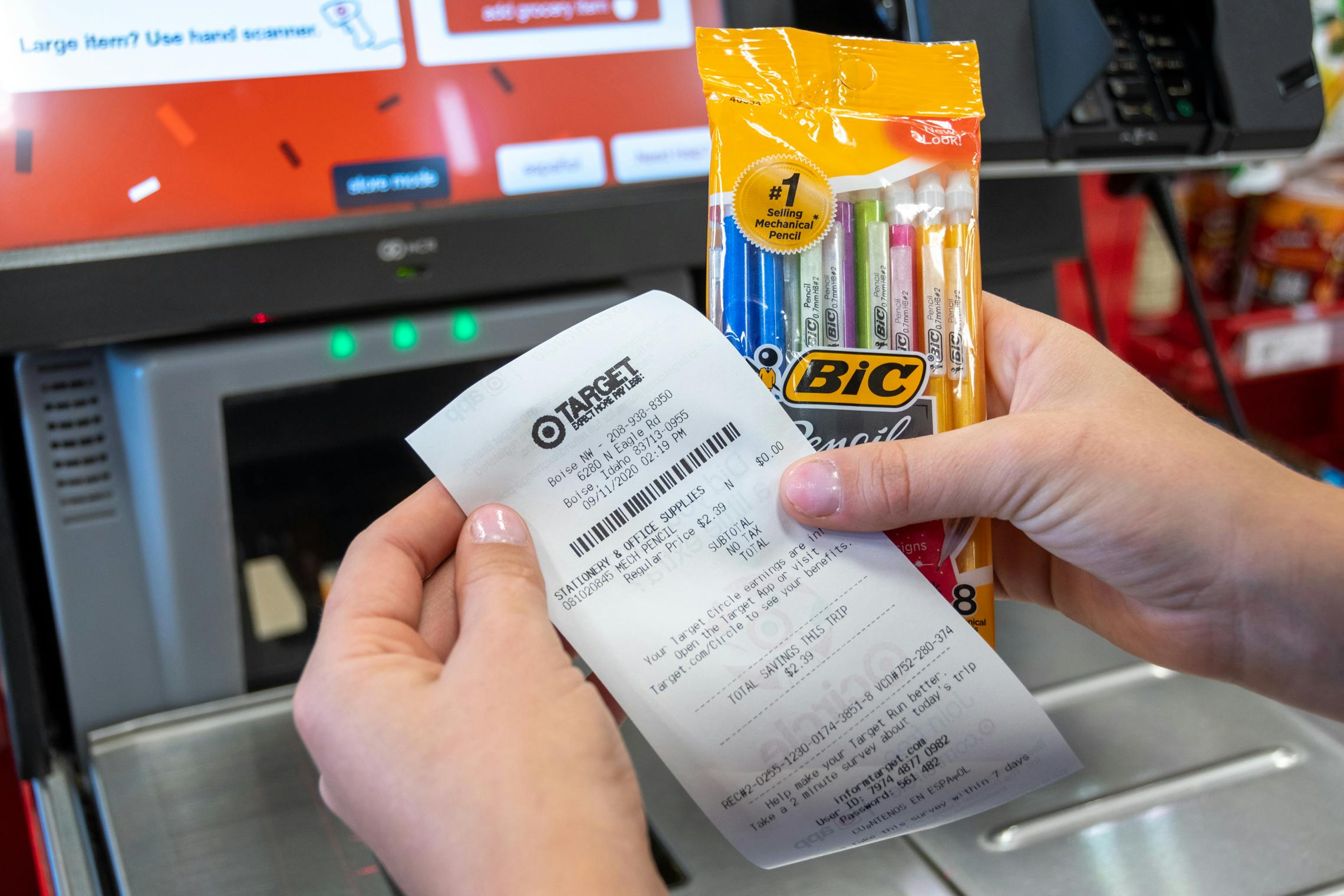 A person at the self-checkout scanner at Target, holding a receipt showing $0 balance next to a pack of BIC mechanical pencils.