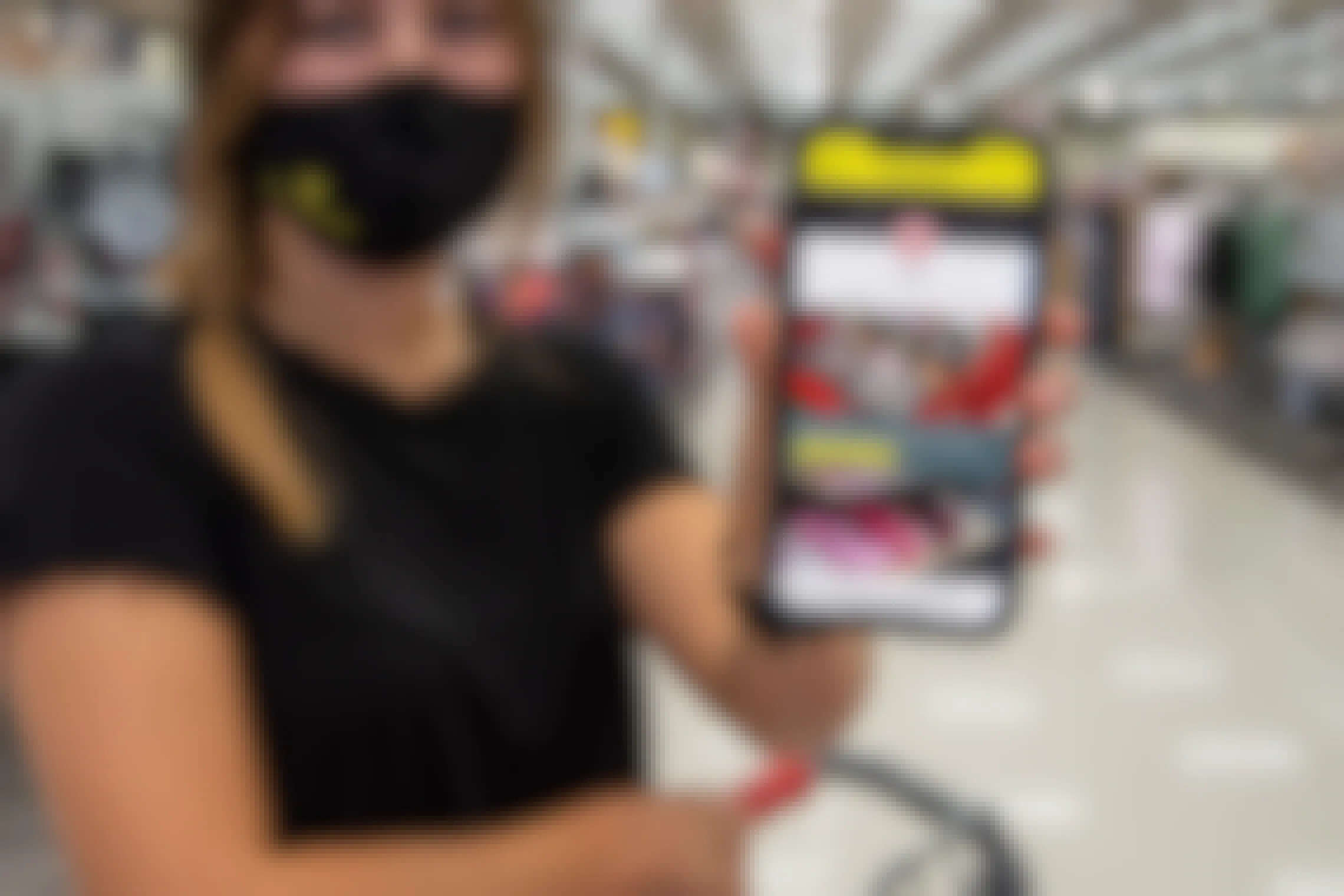 A woman wearing a mask, holding up a phone displaying the KCL app's Target page.