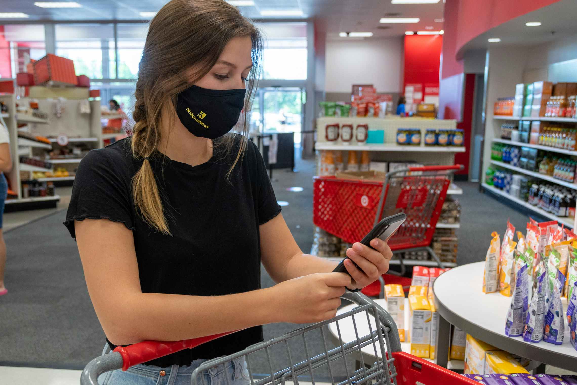 A woman wearing a face mask, standing behind a shopping cart, looking at a cell phone in Target.