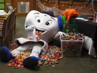 A tooth mascot in a pile of Halloween candy next to a tub filled with more candy.