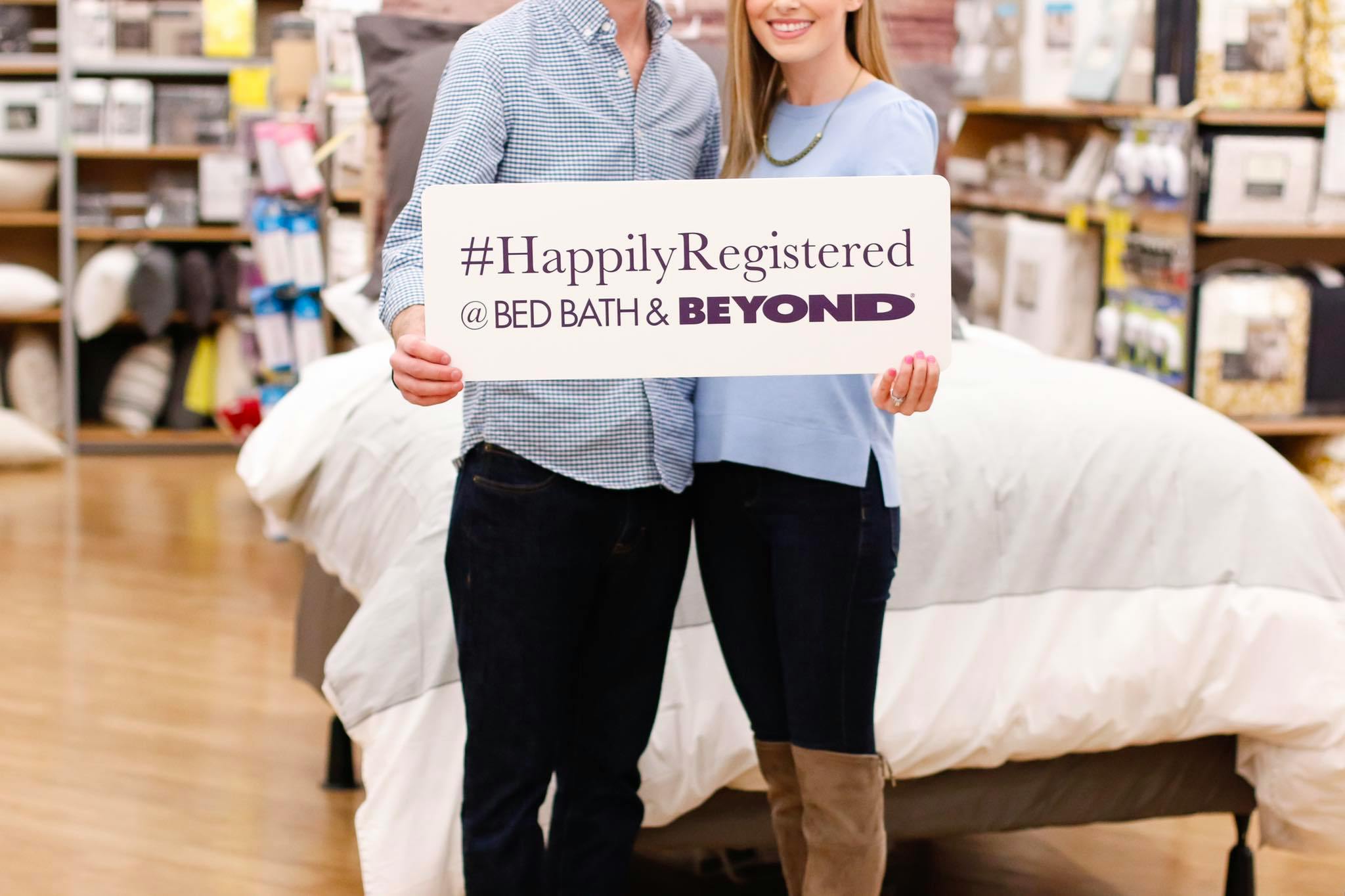 A couple standing inside a Bed Bath & Beyond store, holding a sign that reads, "#HappilyRegistered at Bed Bath & Beyond