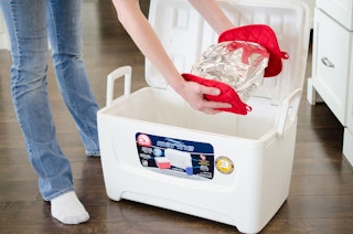 A person placing a food container covered with foil into a large cooler on the floor.