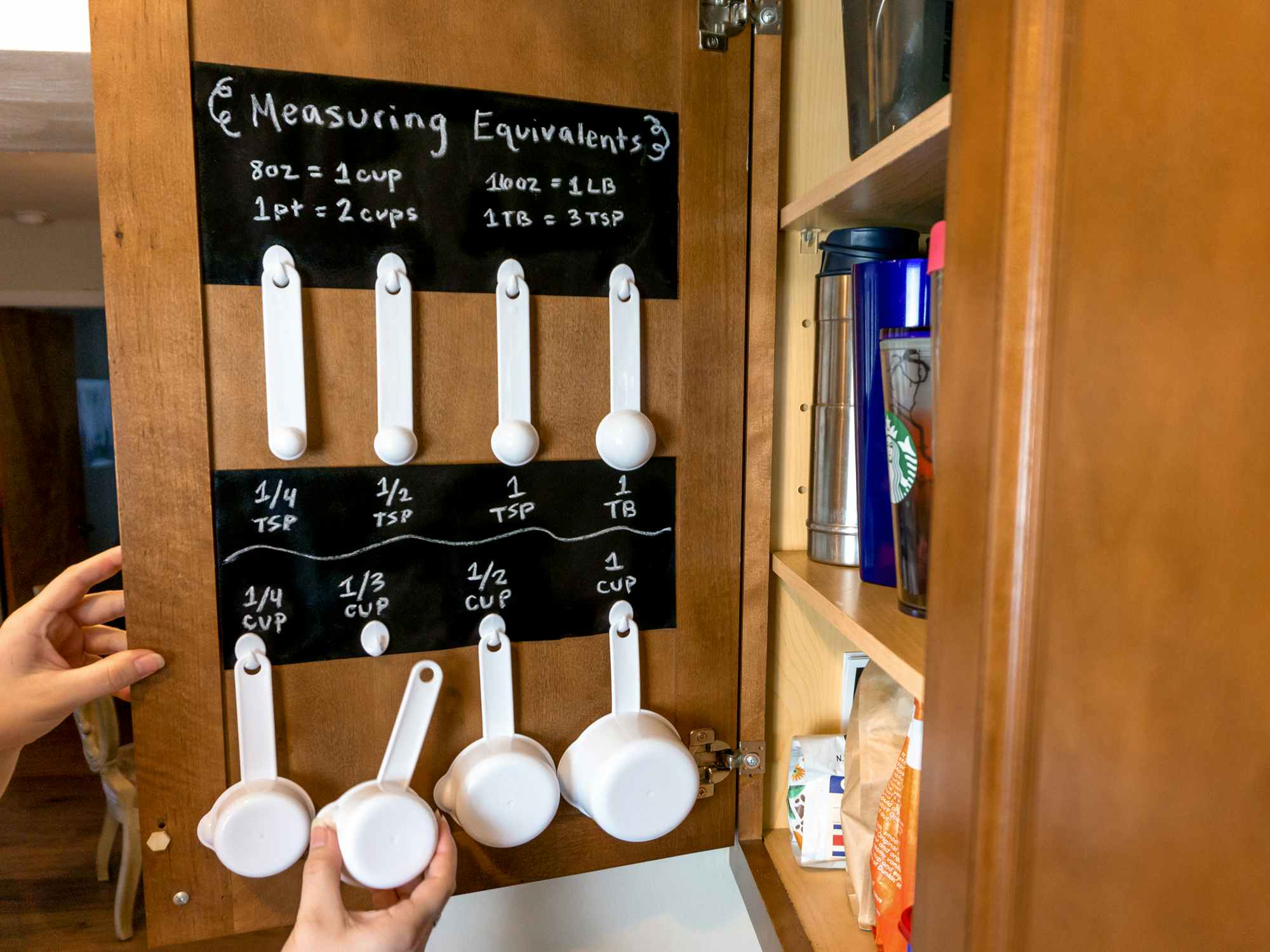 Someone hanging a measuring cup on a hook inside of a kitchen cabinet with chalkboard stickers showing the measurements and common equivalencies