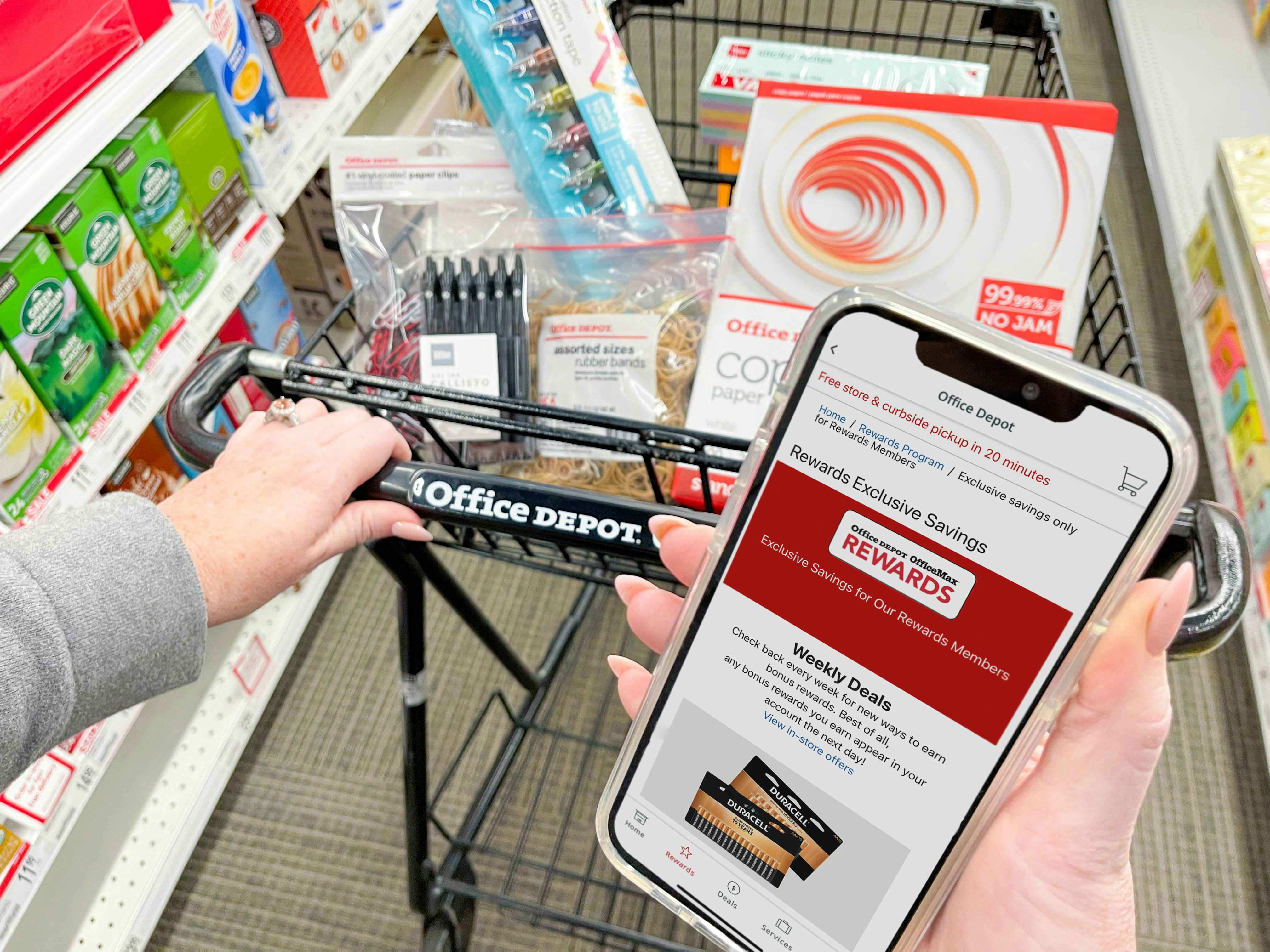 A person's hand holding an iPhone that is displaying the Office Depot mobile app's Rewards page. In the background, the person's other hand is pushing an Office Depot shopping cart full of office supplies down an aisle at Office Depot.