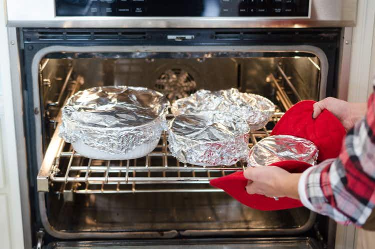 A person taking a foil covered pan out of an oven filled with different sized pans covered with foil.