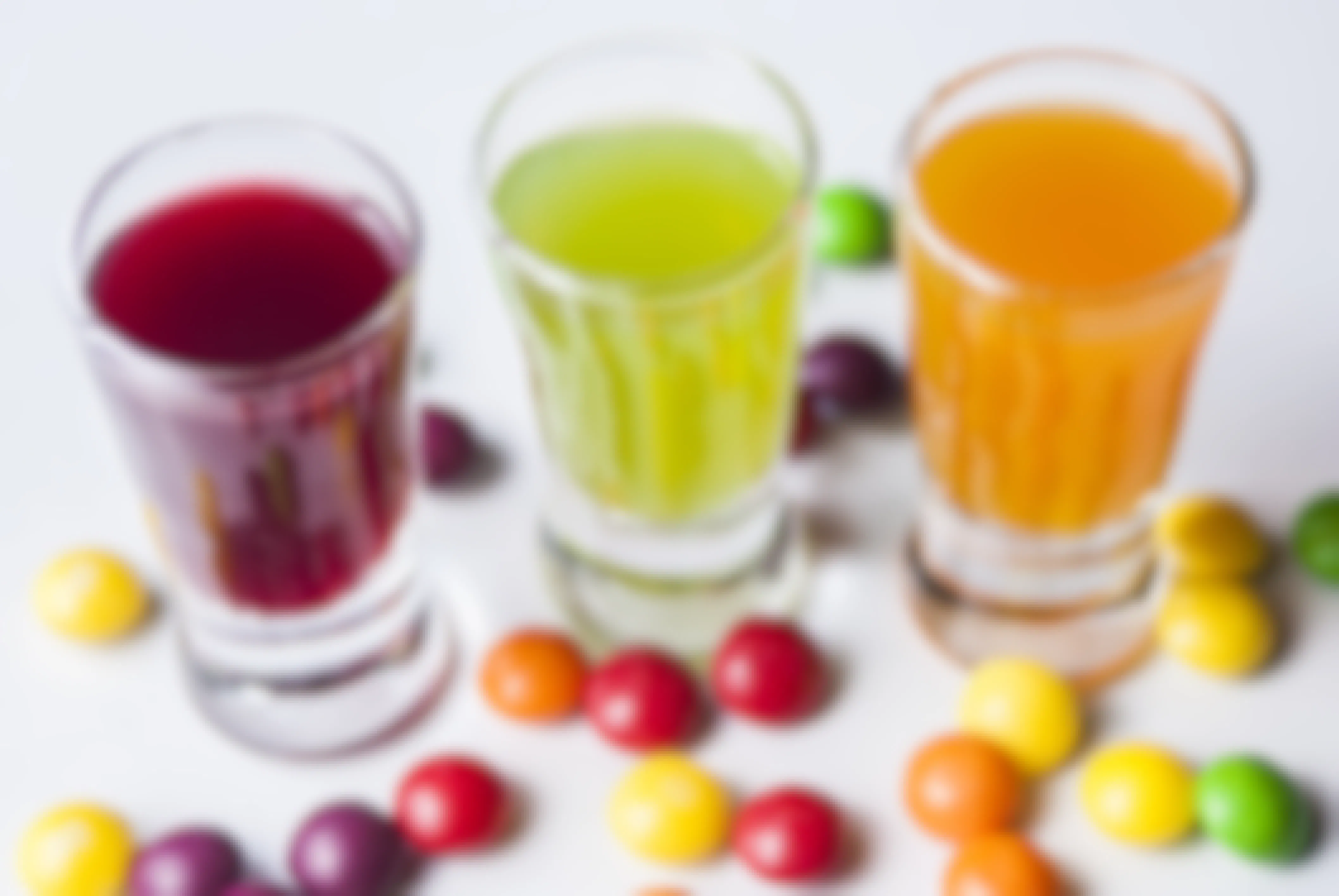 Three shot glasses filled with colorful liquor with colorful candies scattered on the table.