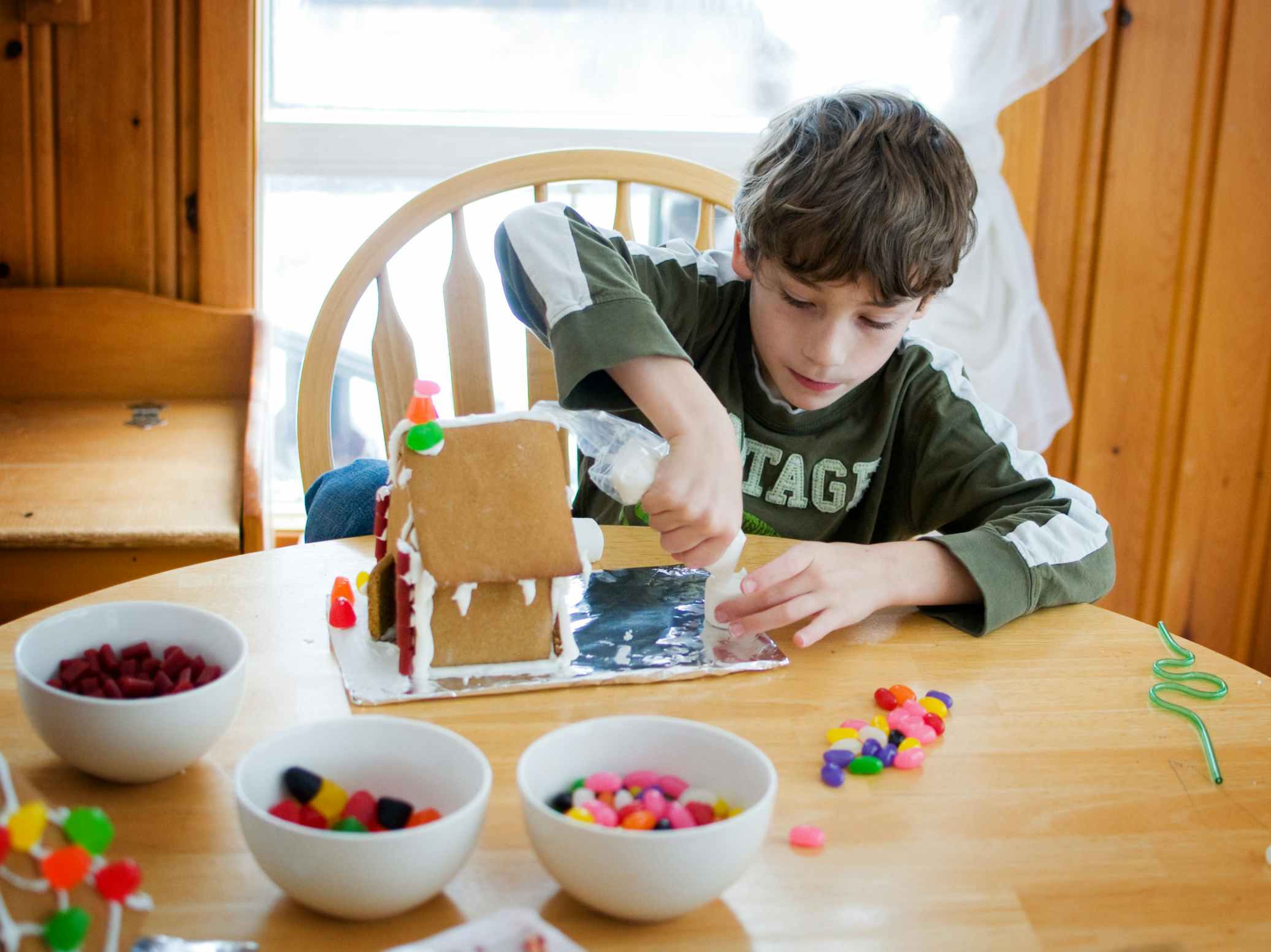A young boy working on decorating a gingerbread house with bowls of different candies on the table.