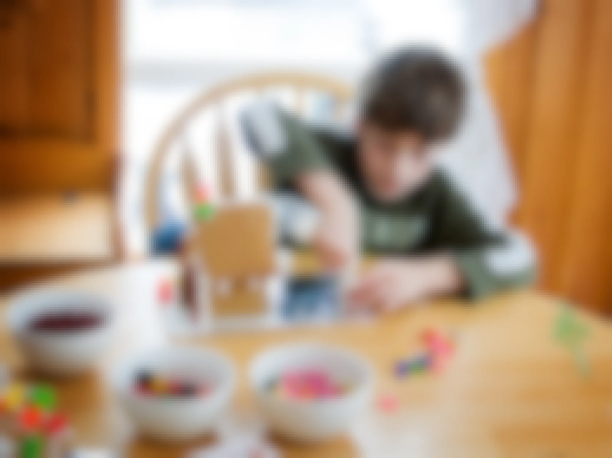 A young boy working on decorating a gingerbread house with bowls of different candies on the table.