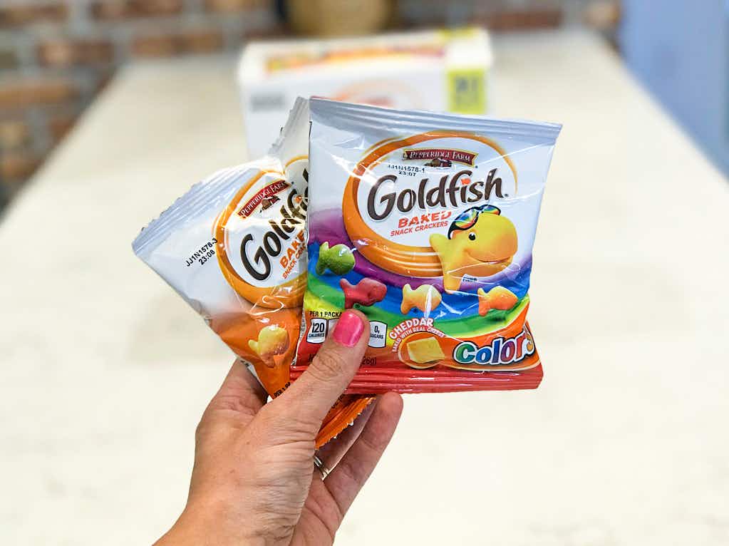 A person's hand holding up two snack packs of Goldfish crackers in front of a counter.