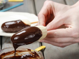 A person dipping a banana half on a popsicle stick into some melted chocolate.
