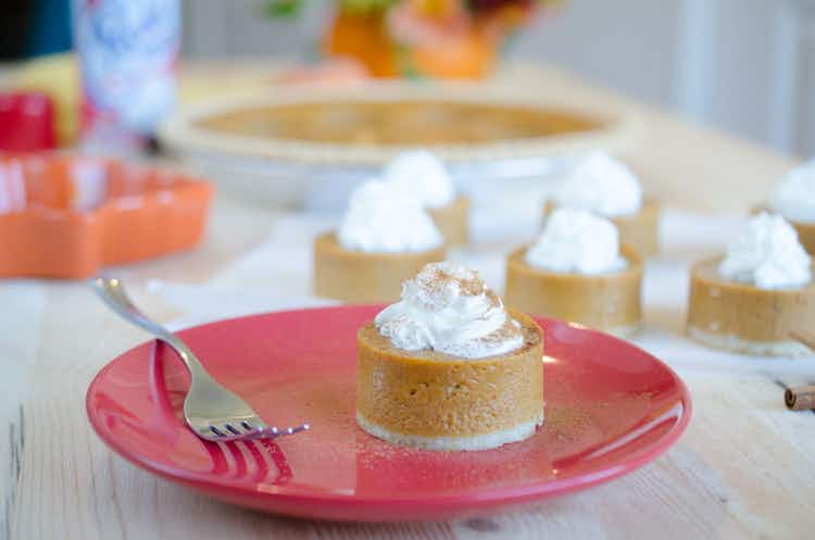 A mini pumpkin pie with whipped cream sitting on a plate in front of more of the same dessert on the counter.