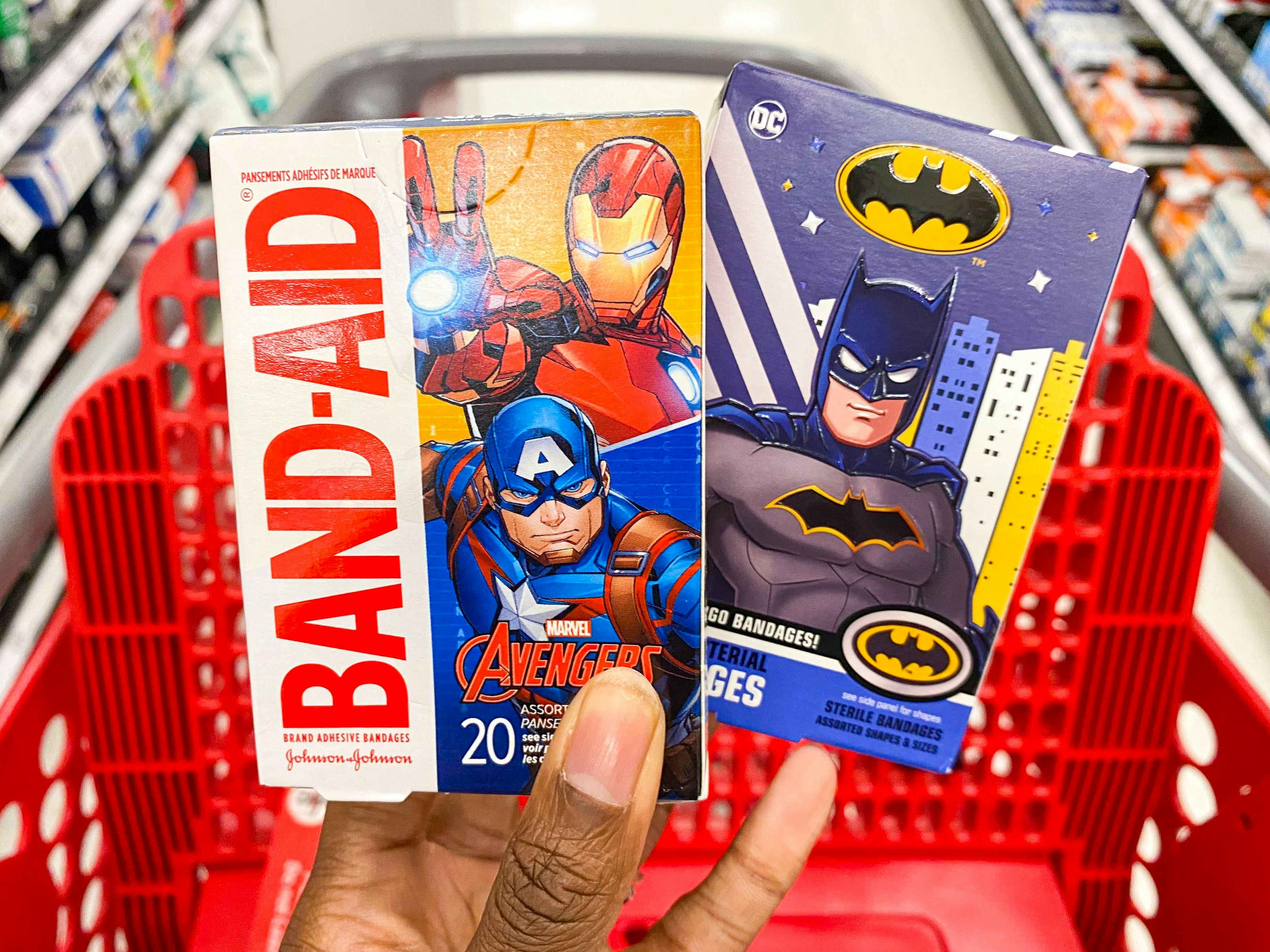 A person's hand holding two boxes of superhero Band-Aids in front of a Target shopping cart.