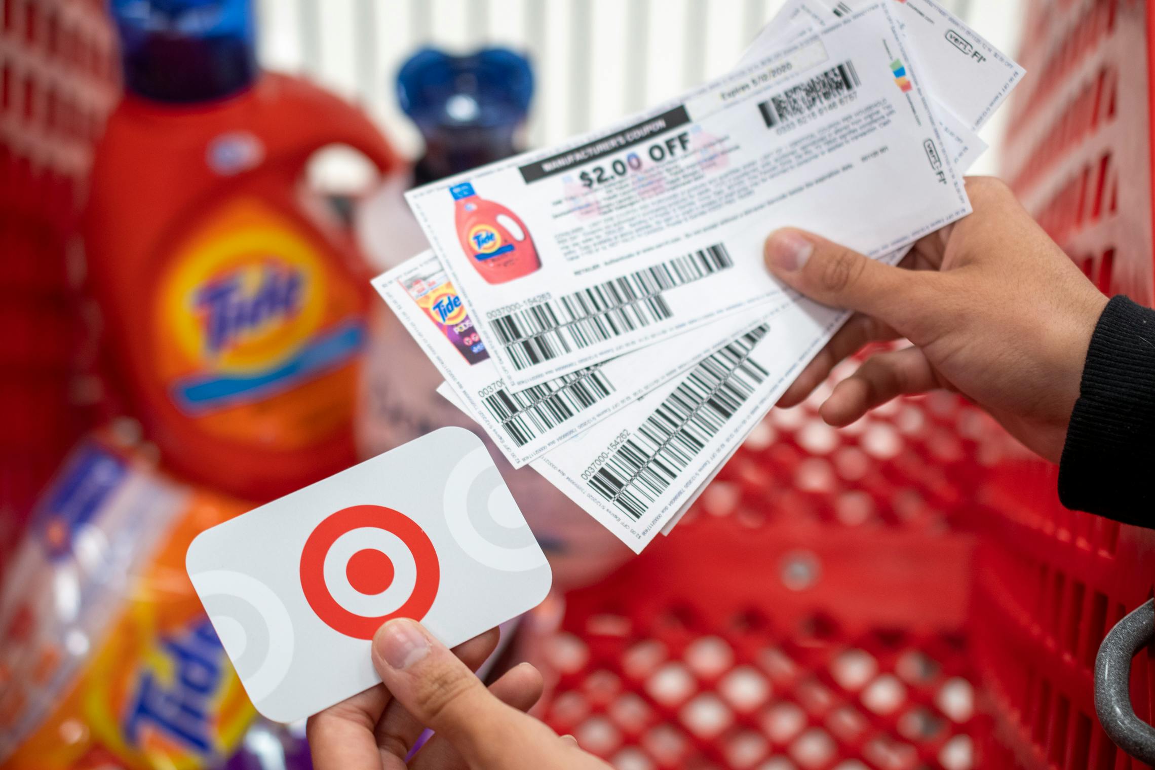 A person holding tide manufacture coupons in one hand and a Target gift card in the other, with a shopping card containing laundry detergents in the background.
