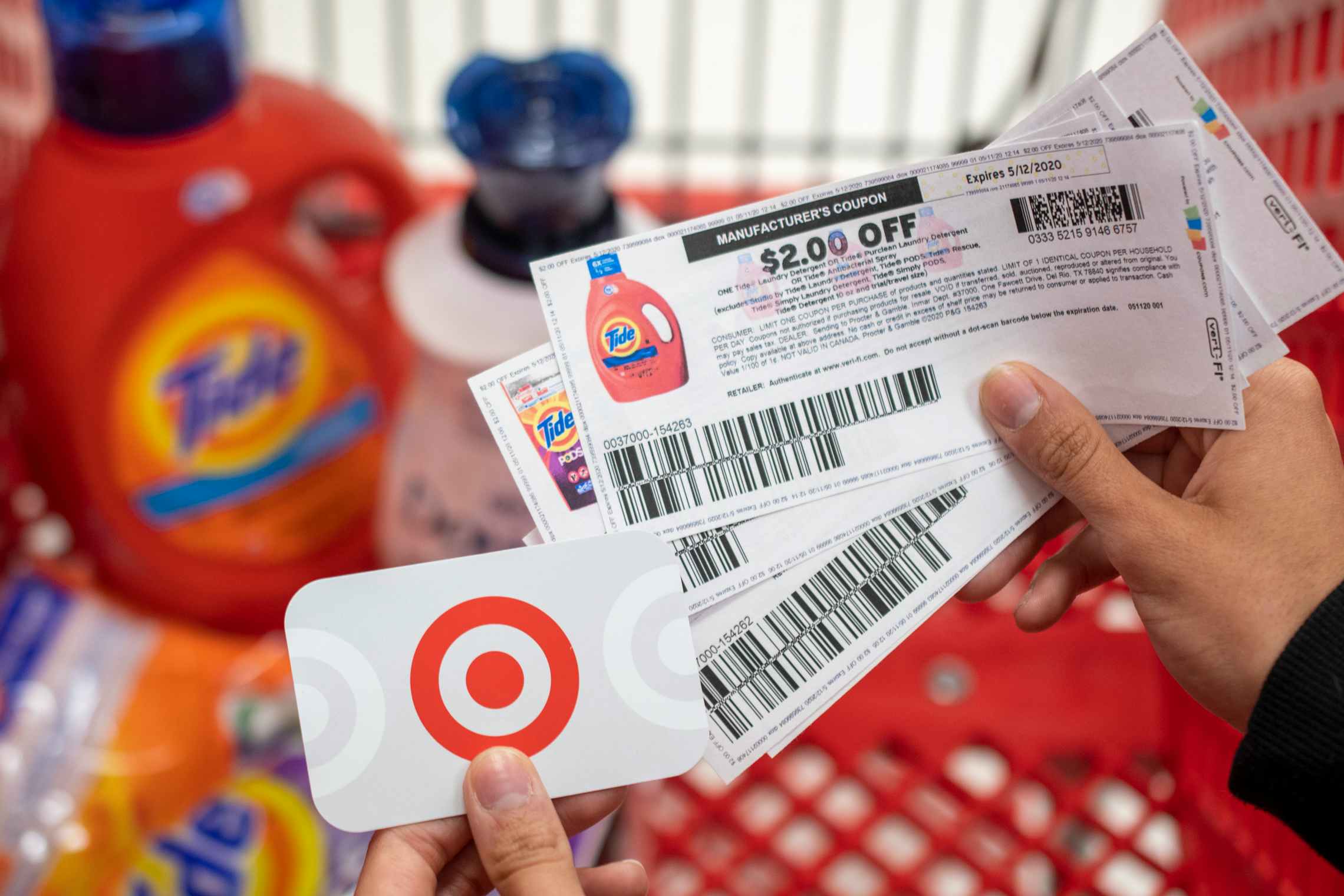 A person holding tide manufacture coupons in one hand and a Target gift card in the other, with a shopping card containing laundry detergents in the background.