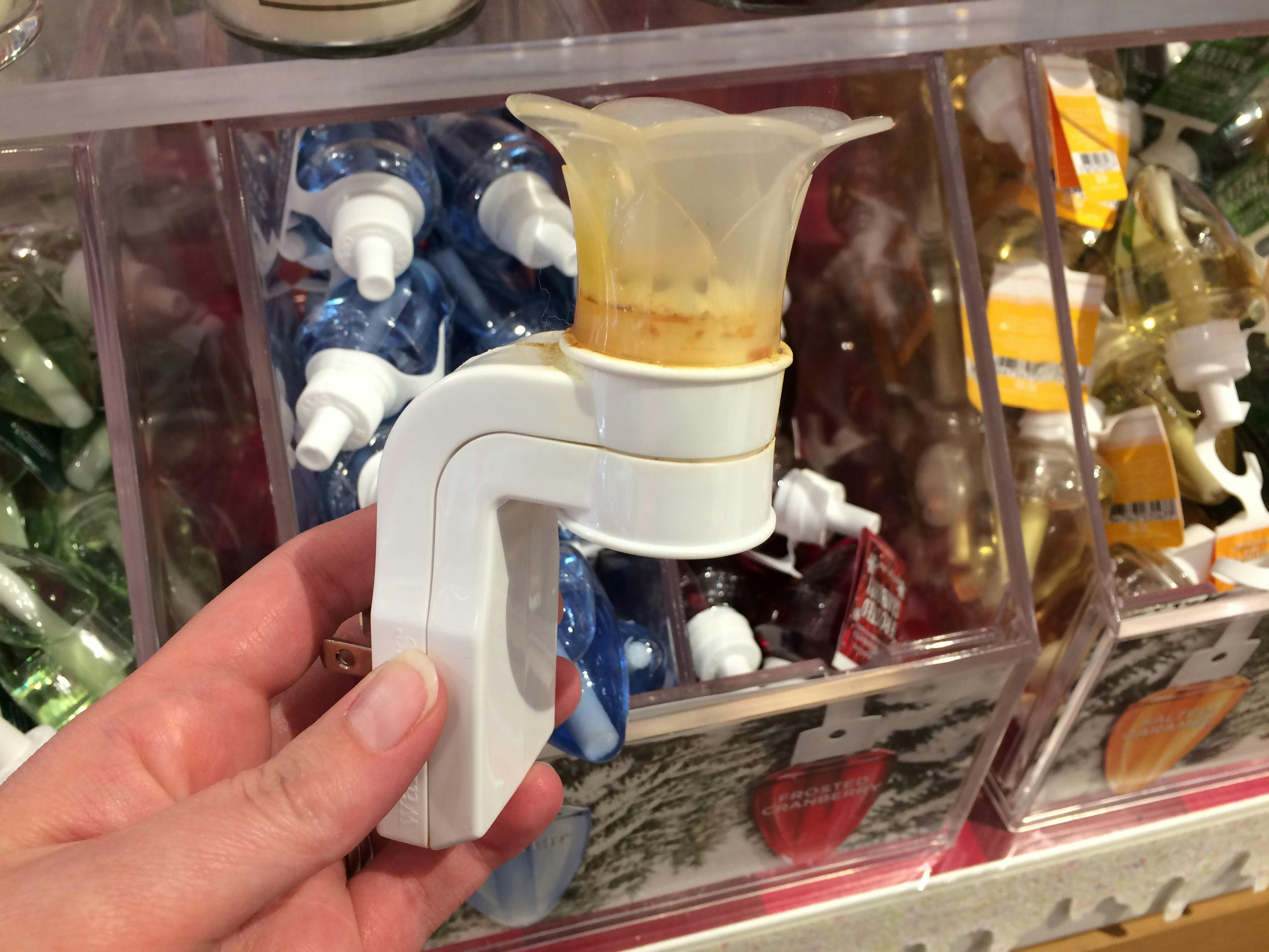 A person's hand holding a used Wallflower plugin in front of bins filled with Wallflower refills at Bath & Body Works..