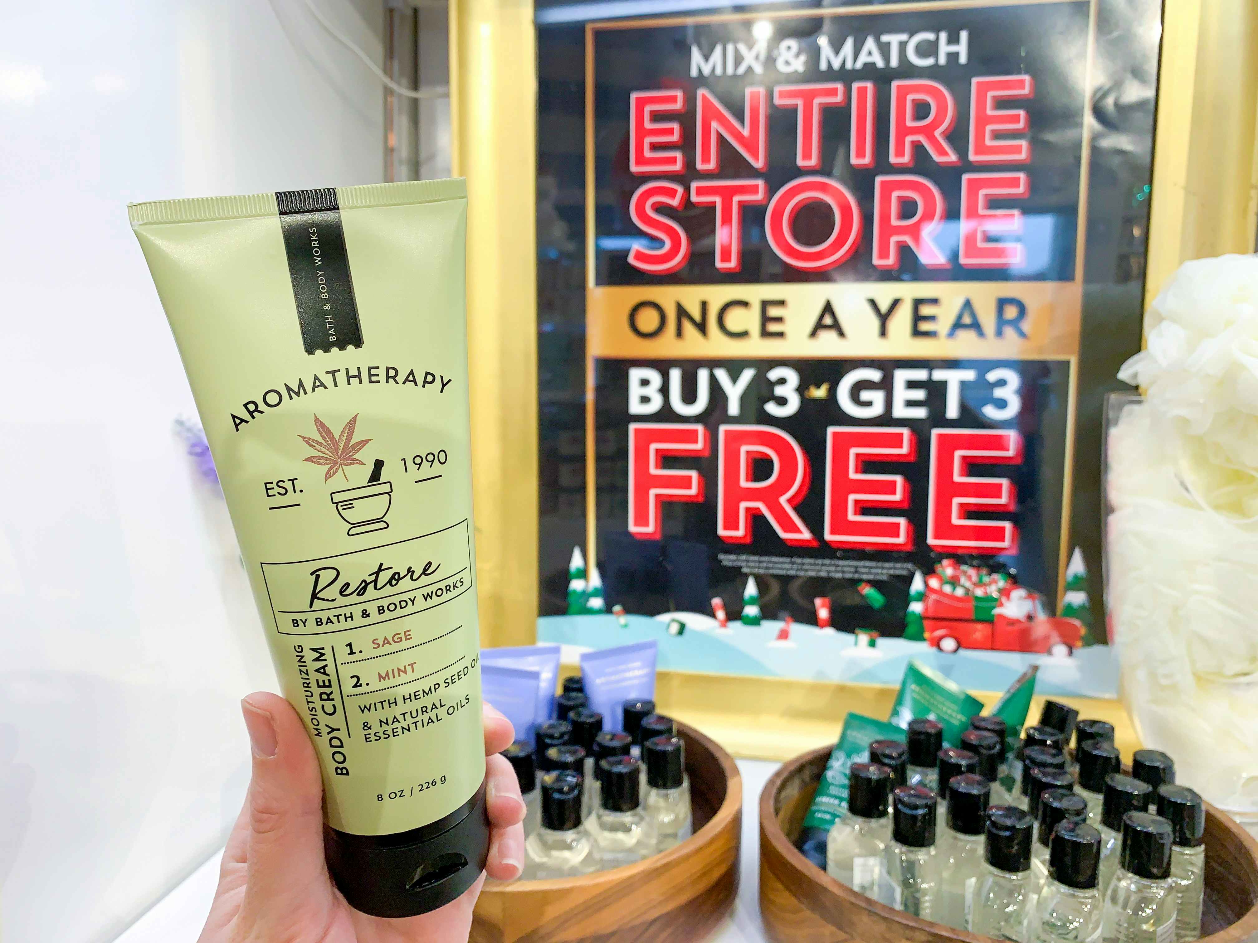 A person's hand holding up a bottle of Aromatherapy lotion in front of a big sign inside Bath & Body Works that reads, "Mix & Match, entire store, once a year, buy 3 get 3 free