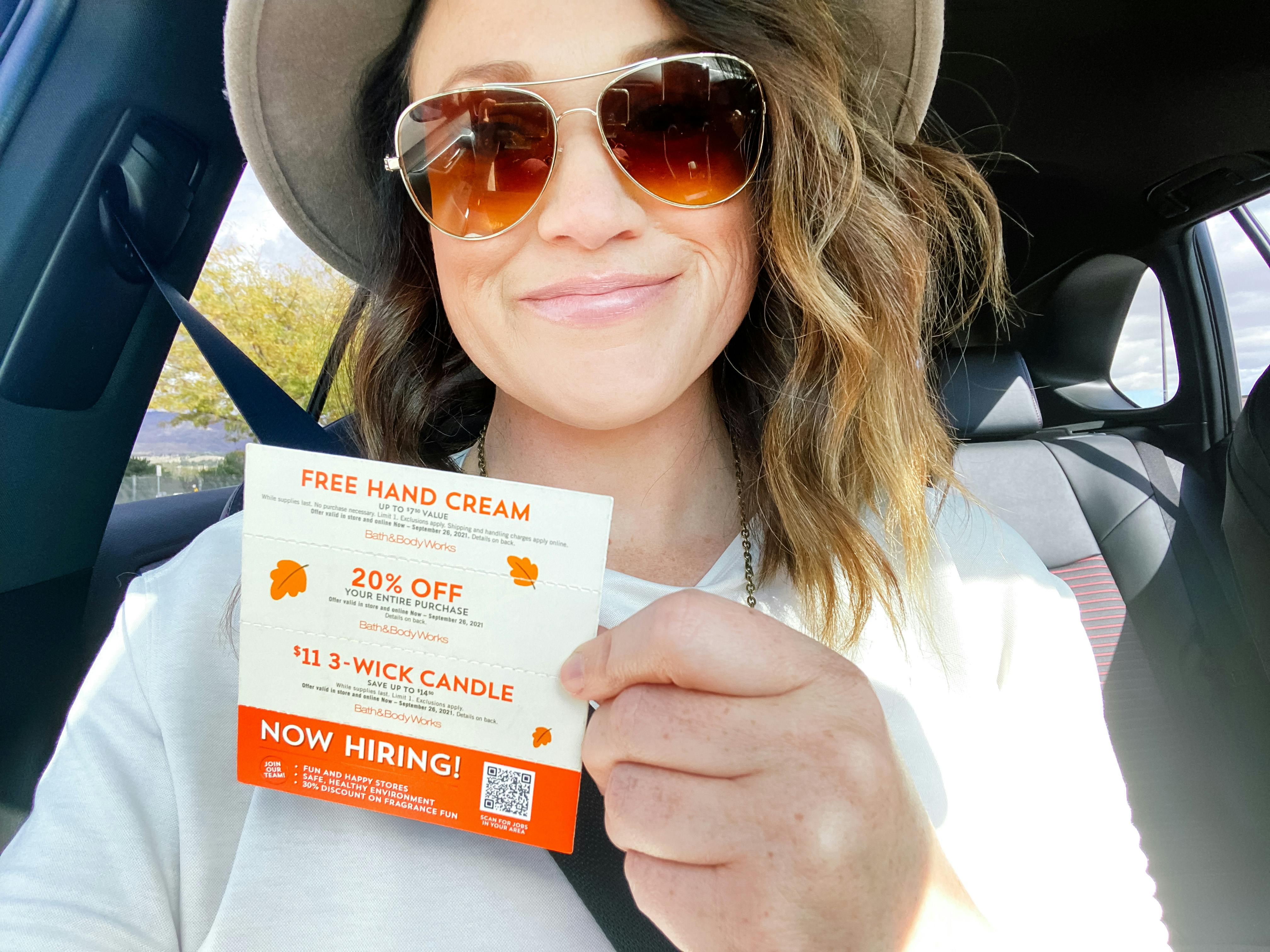 A woman sitting in her car, smiling, holding up a Bath & Body Works coupon mailer with multiple coupons on it.