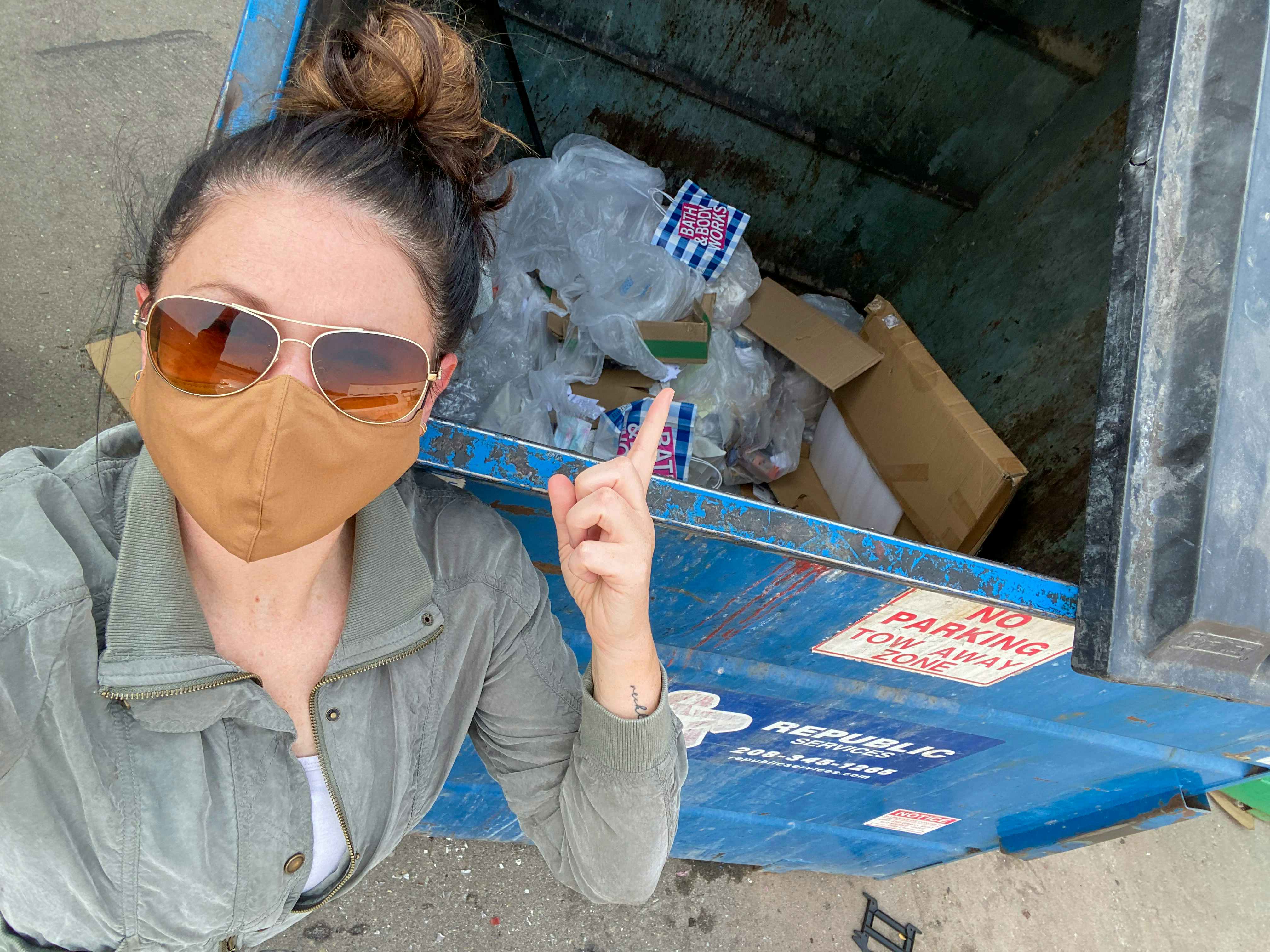 A woman standing by a dumpster, pointing to the discarded Bath & Body Works bags and products inside.