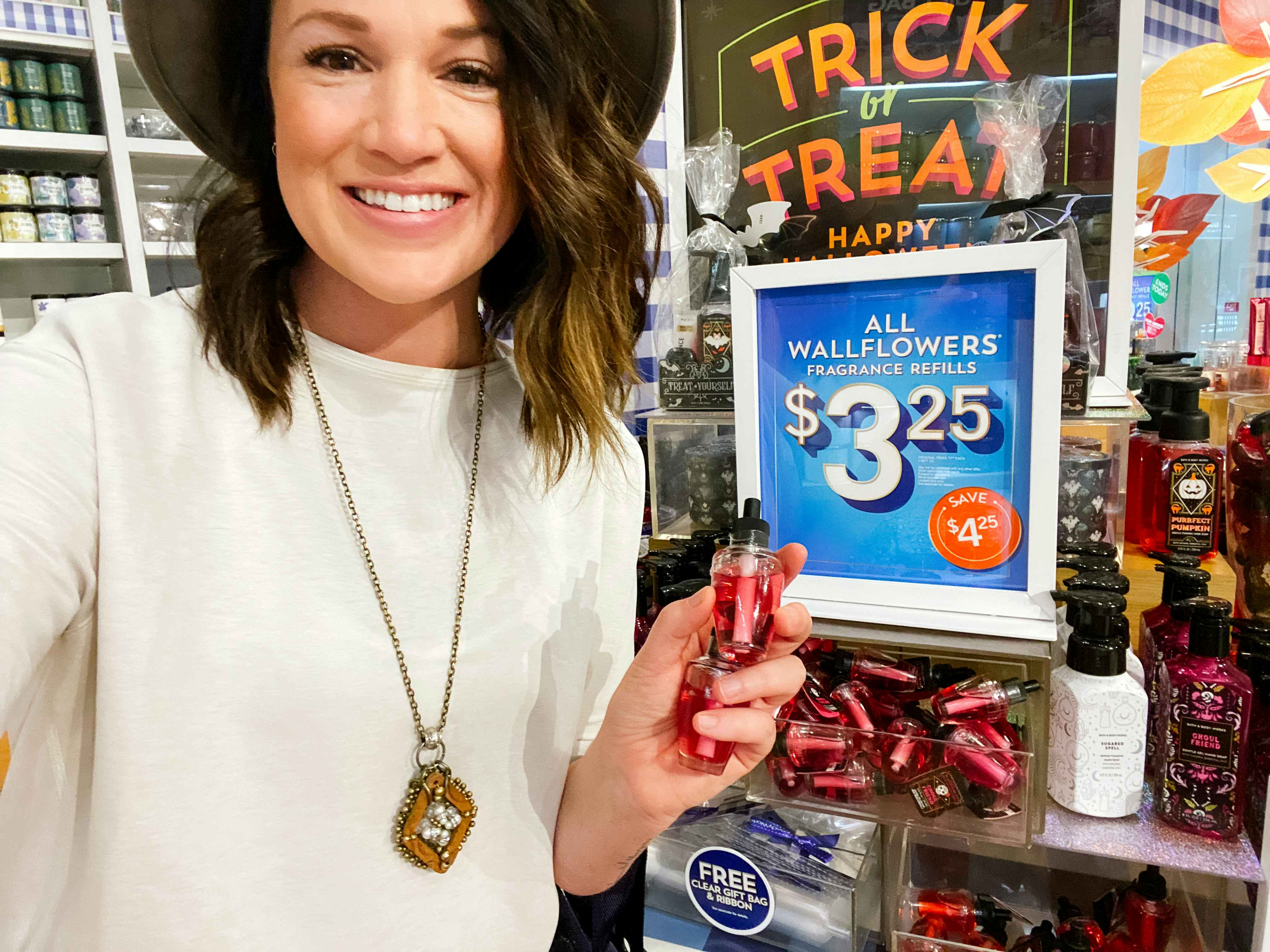 A woman smiling and holding up a Bath & Body Works wallflower refill in front of a sign that reads, "All wallflowers fragrance refills $3.25" inside Bath & Body Works.