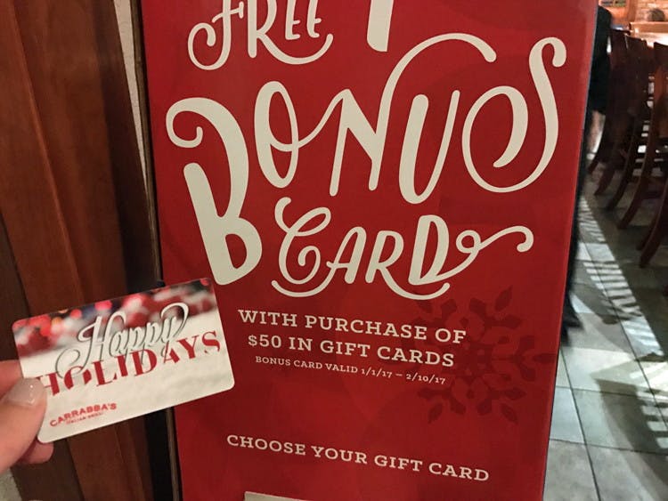 someone holding carrabbas gift card in front of a sign that says free bonus card with purchase of $50 in gift cards