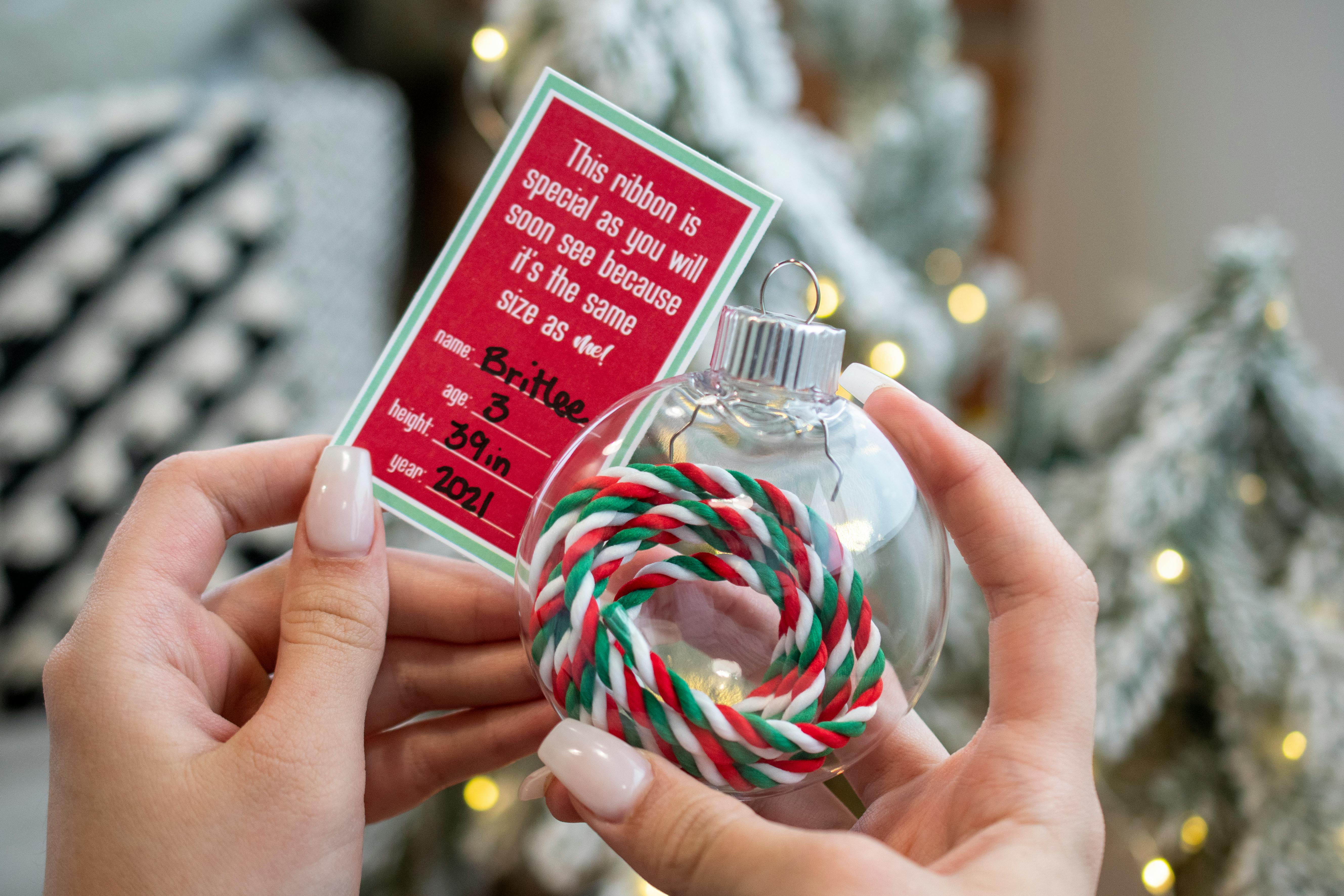 Someone holding a clear ornament with a ribbon inside it with a tag that says "This ribbon is special as you will soon see because it's the same size as me! With the name, age, height of a child and the year